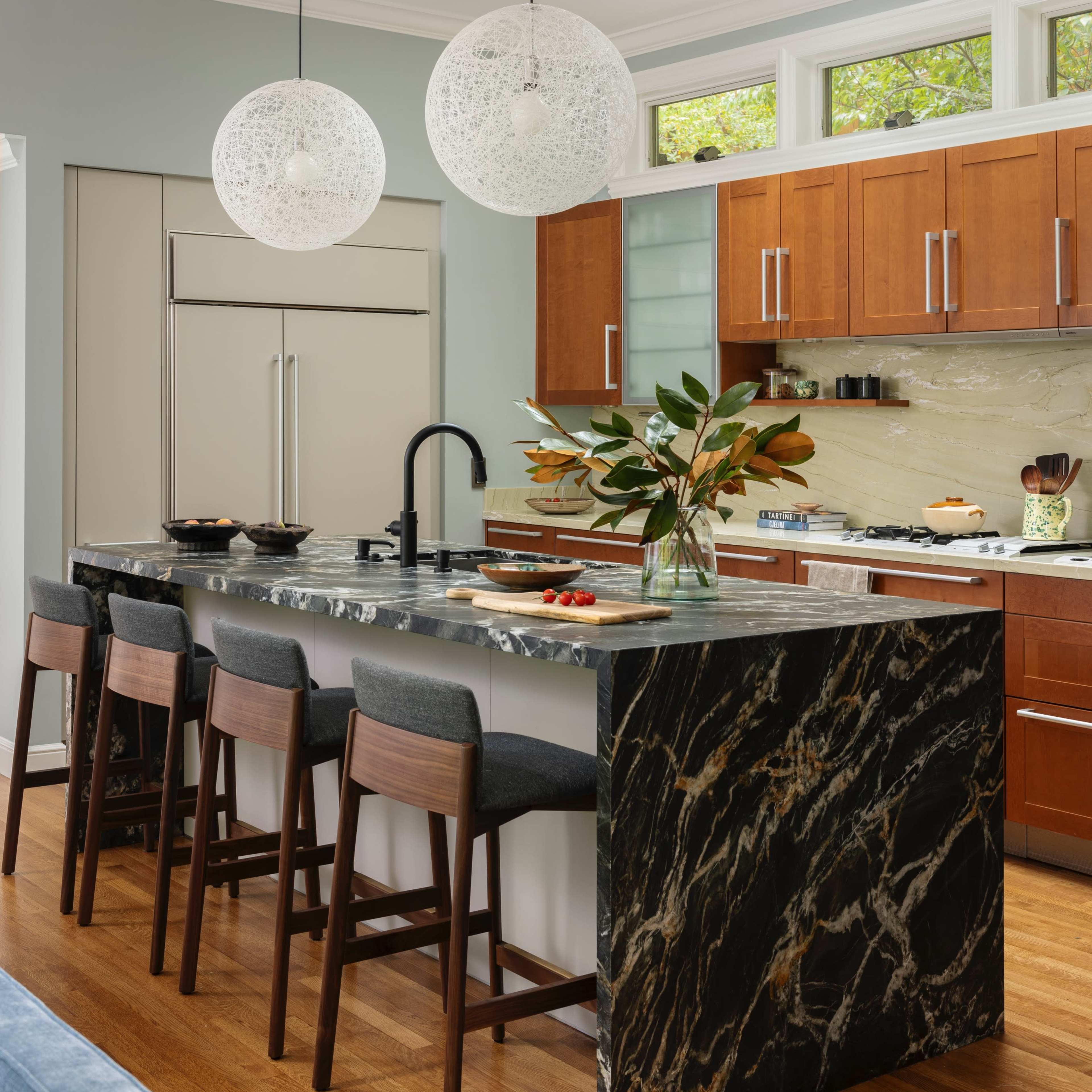 A modern kitchen features a marble island with seating, wooden cabinetry, and pendant lights above the countertop.
