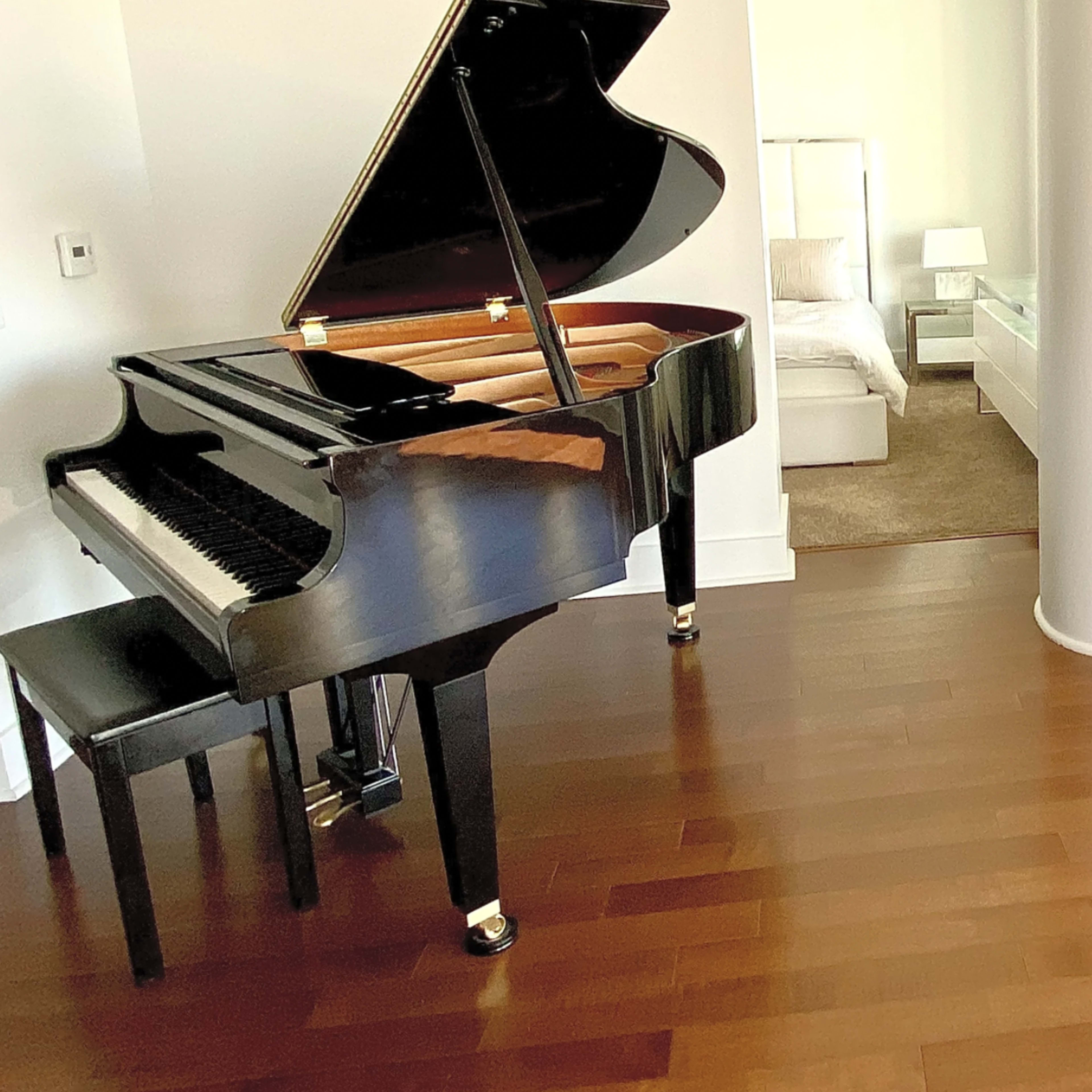 A grand piano sits next to a light-filled bedroom with sliding glass doors and hardwood flooring.