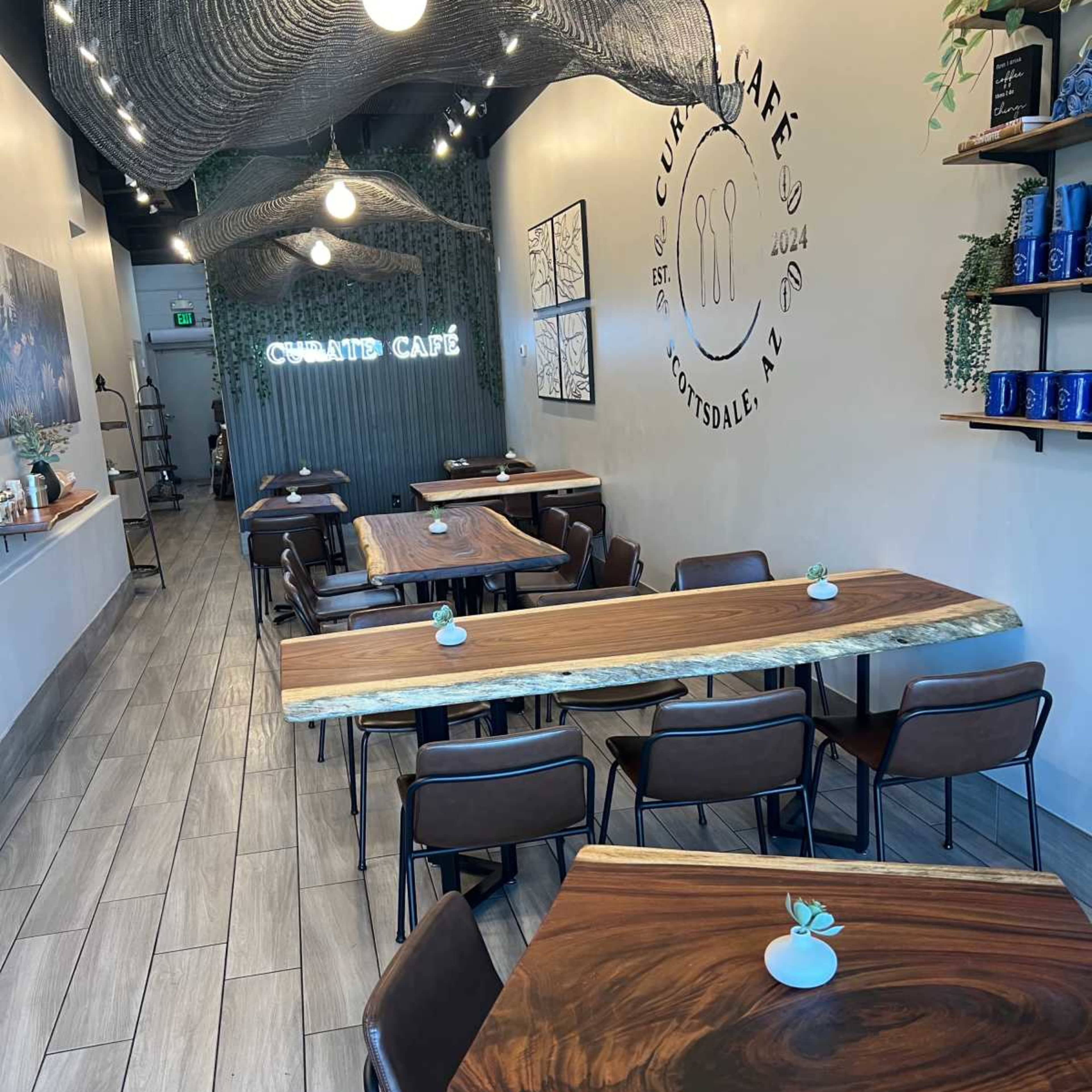 A modern café interior with wooden tables, brown chairs, and a minimalist design featuring a decorative wall with the café's name and plants.