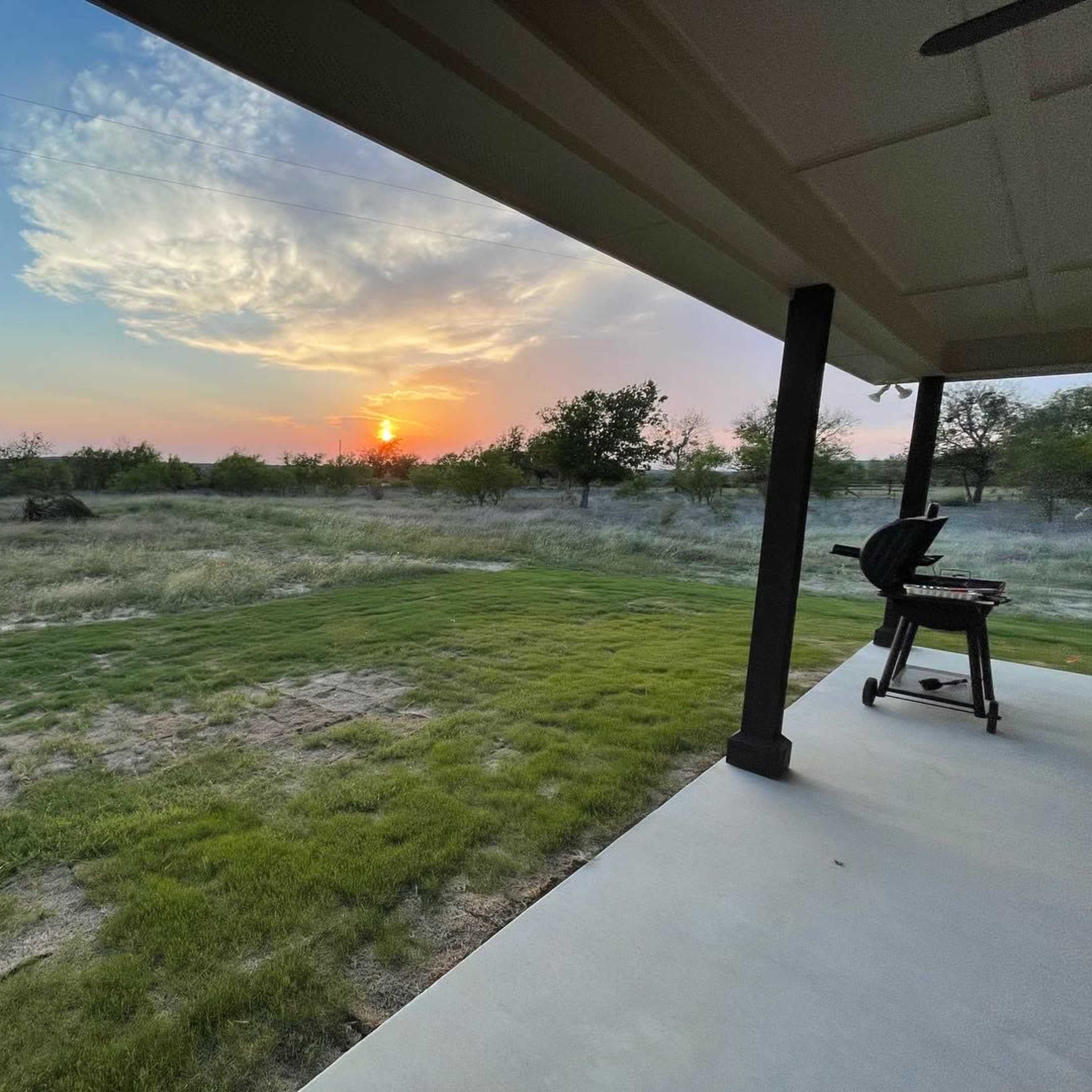 A sunset over a grassy landscape viewed from a covered patio with a grill.