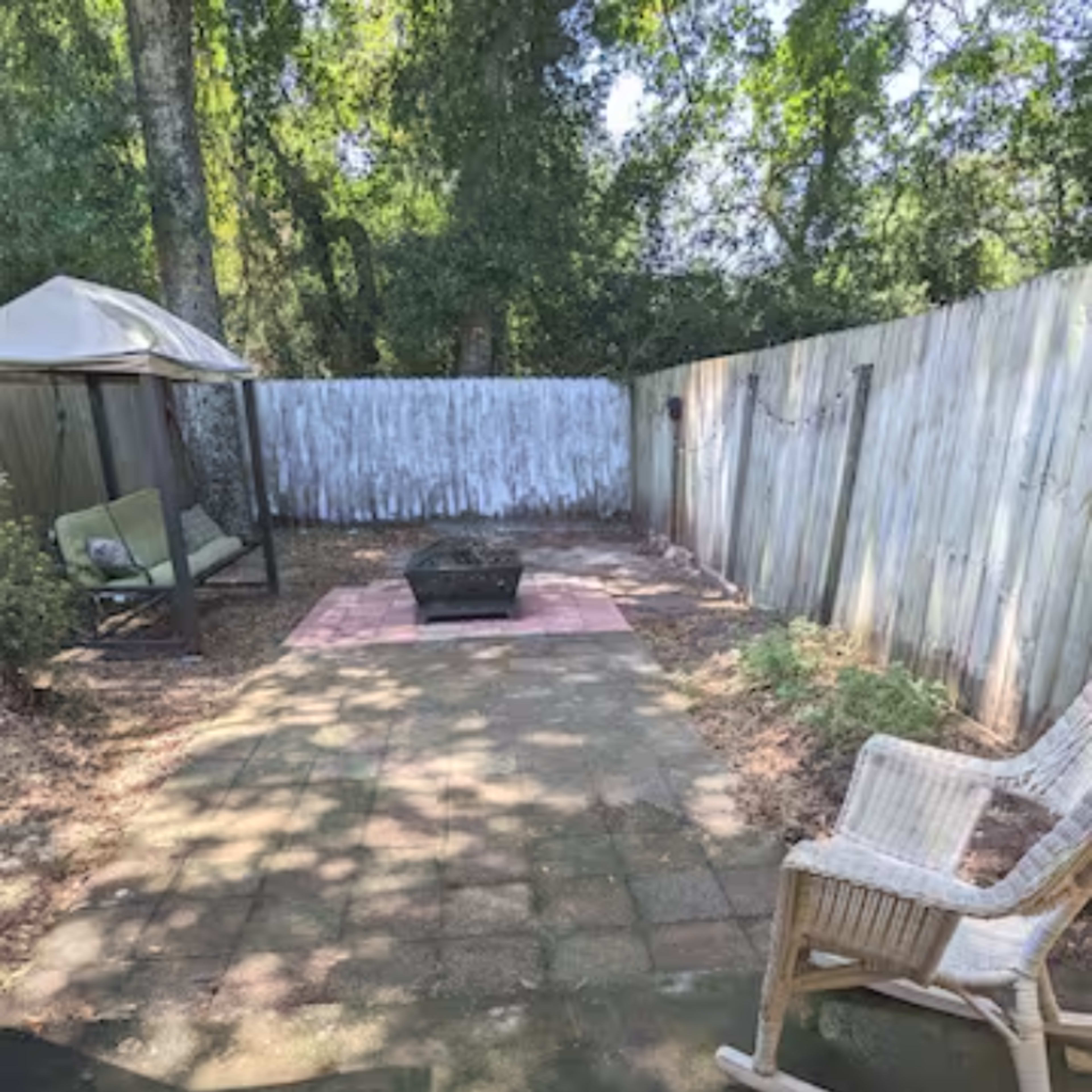 A paved backyard area with a fire pit at the center, surrounded by a gazebo and a rocking chair, with a wooden fence in the background.
