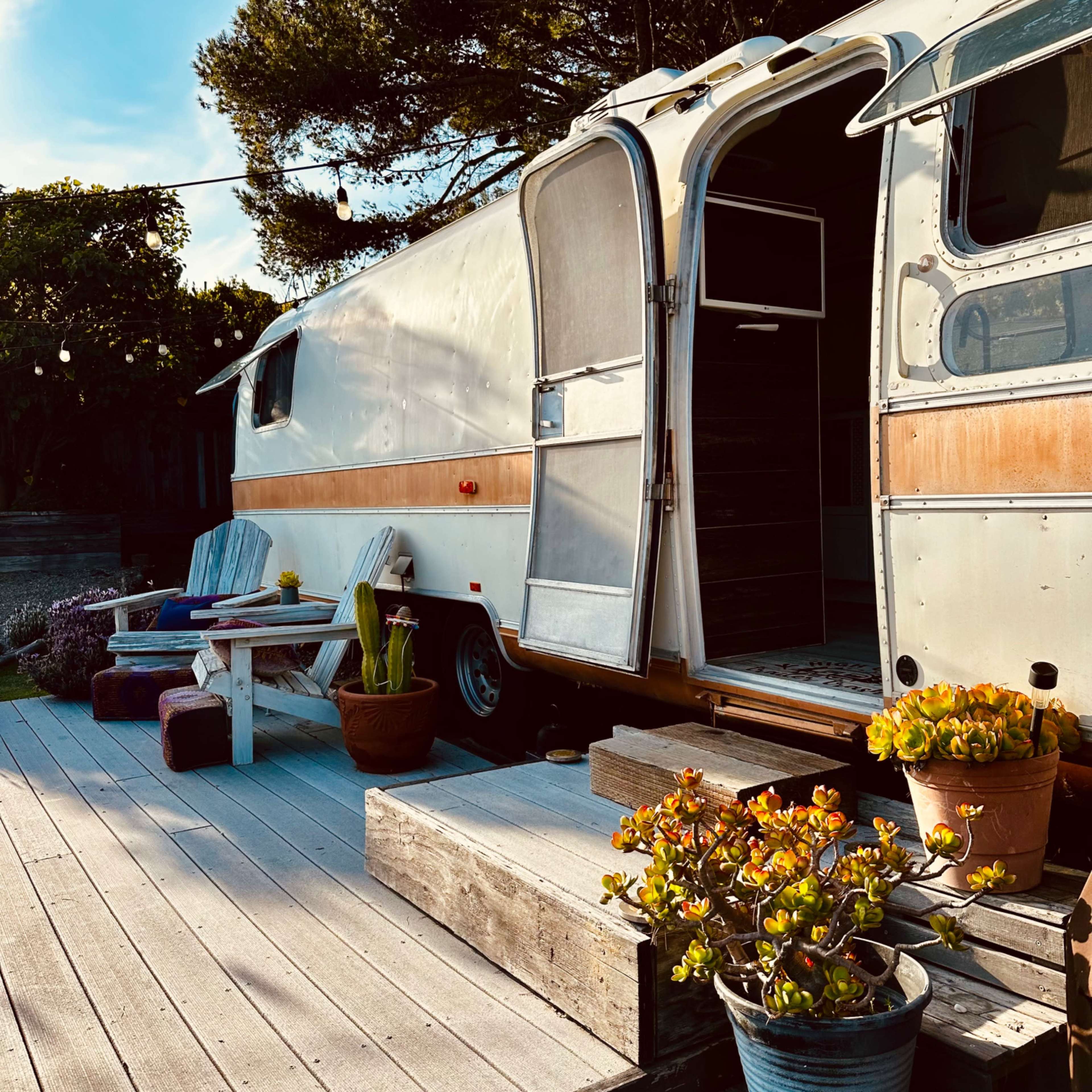 An Airstream trailer is parked next to a wooden deck adorned with potted plants and outdoor seating.