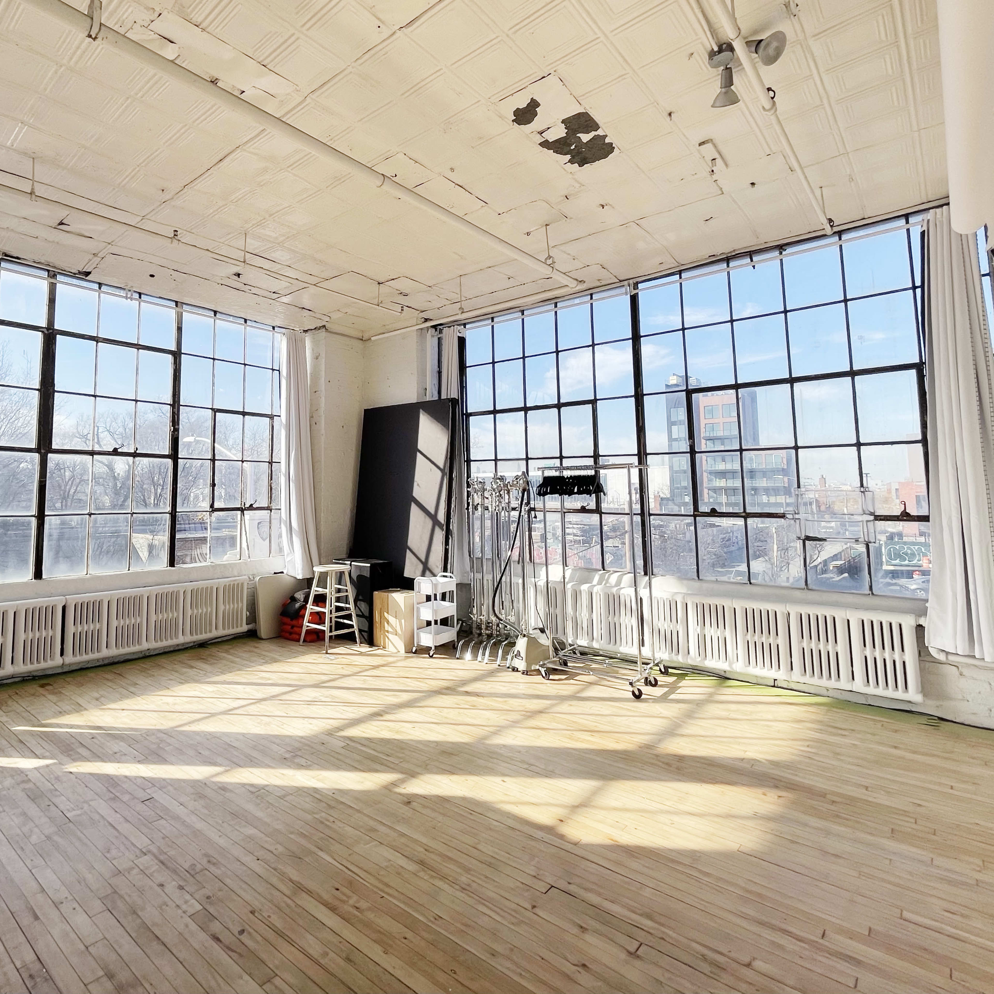 A spacious, sunlit room with large windows, wooden flooring, and minimal furnishings.