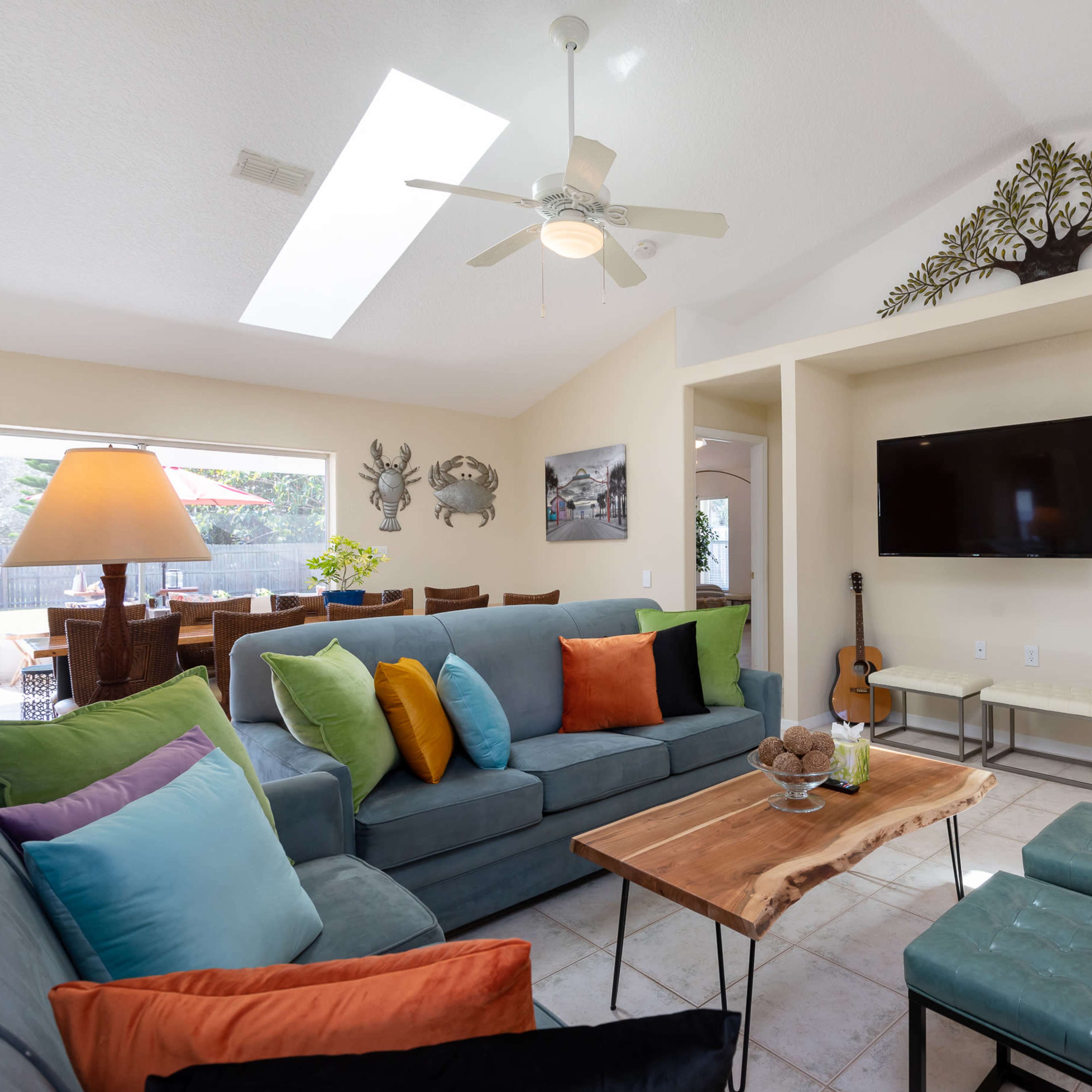 A modern living room featuring a sectional sofa with colorful cushions, a wooden coffee table, and a wall-mounted television, all illuminated by natural light from a skylight.