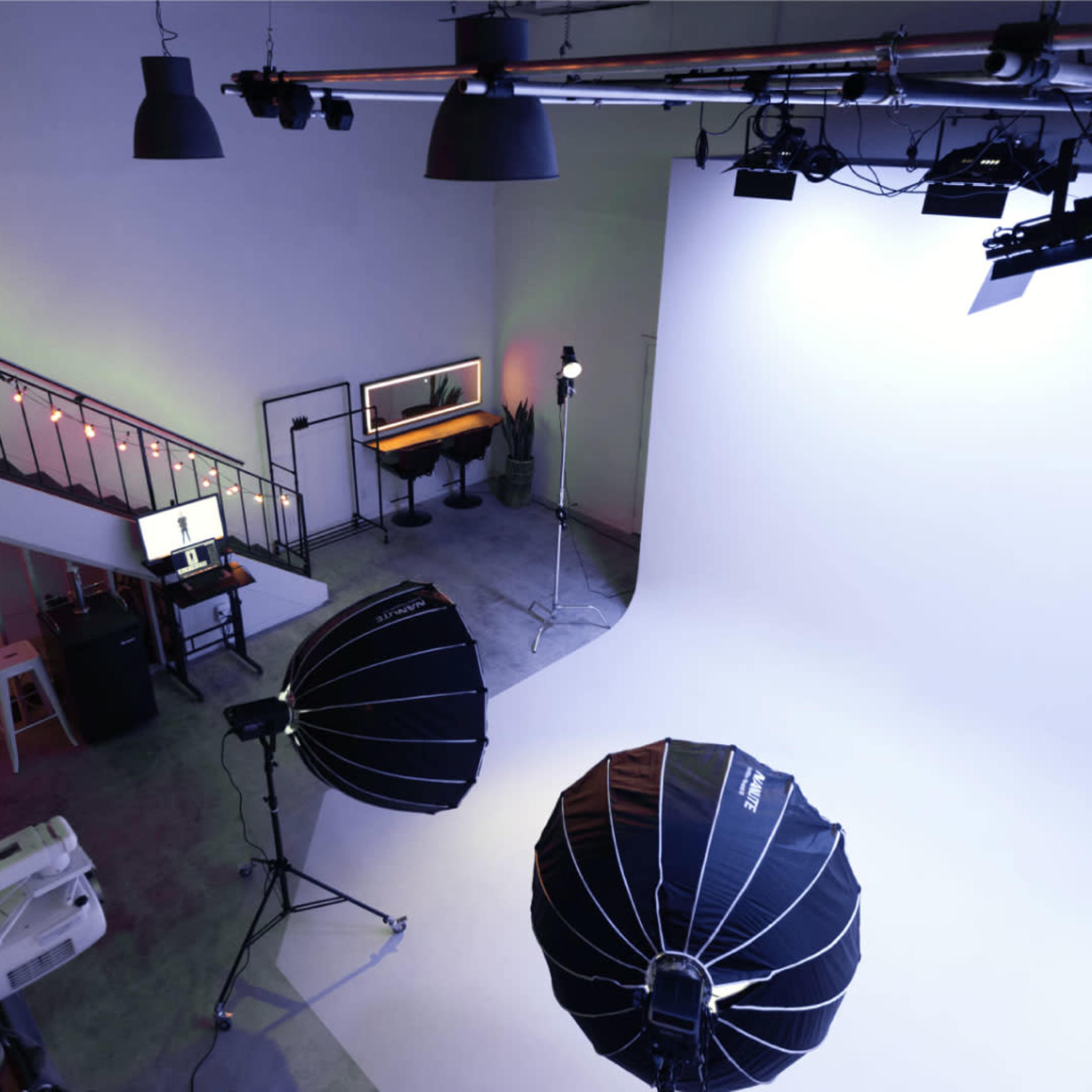 A photography studio features a large white backdrop, lighting equipment, and a workspace with a computer and seating on an upper level.
