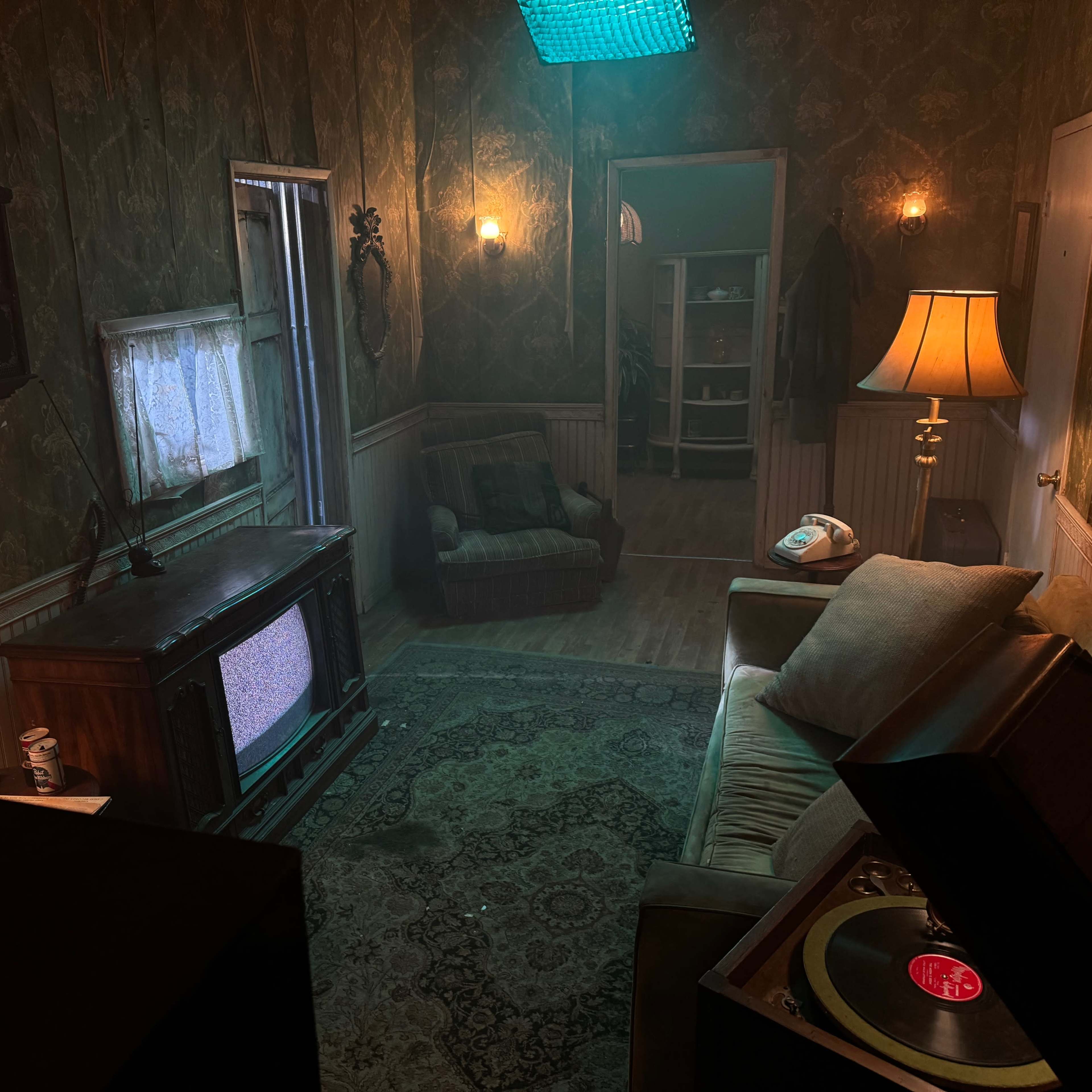A dimly lit room with vintage furniture, including a sofa, a record player, and a television, all set against patterned wallpaper.