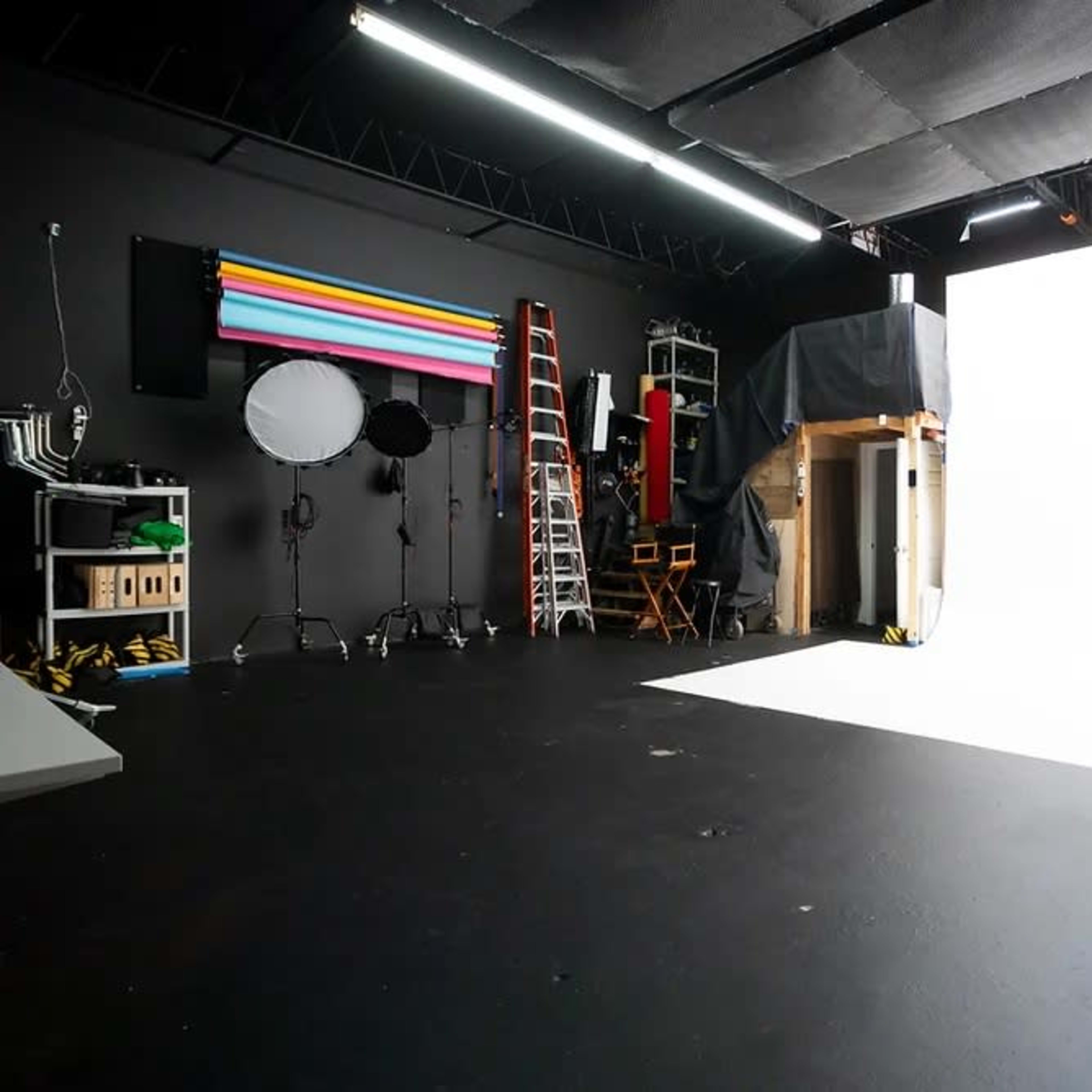An empty photography studio with various equipment, including lighting, backdrops, and a ladder, set against a white backdrop.