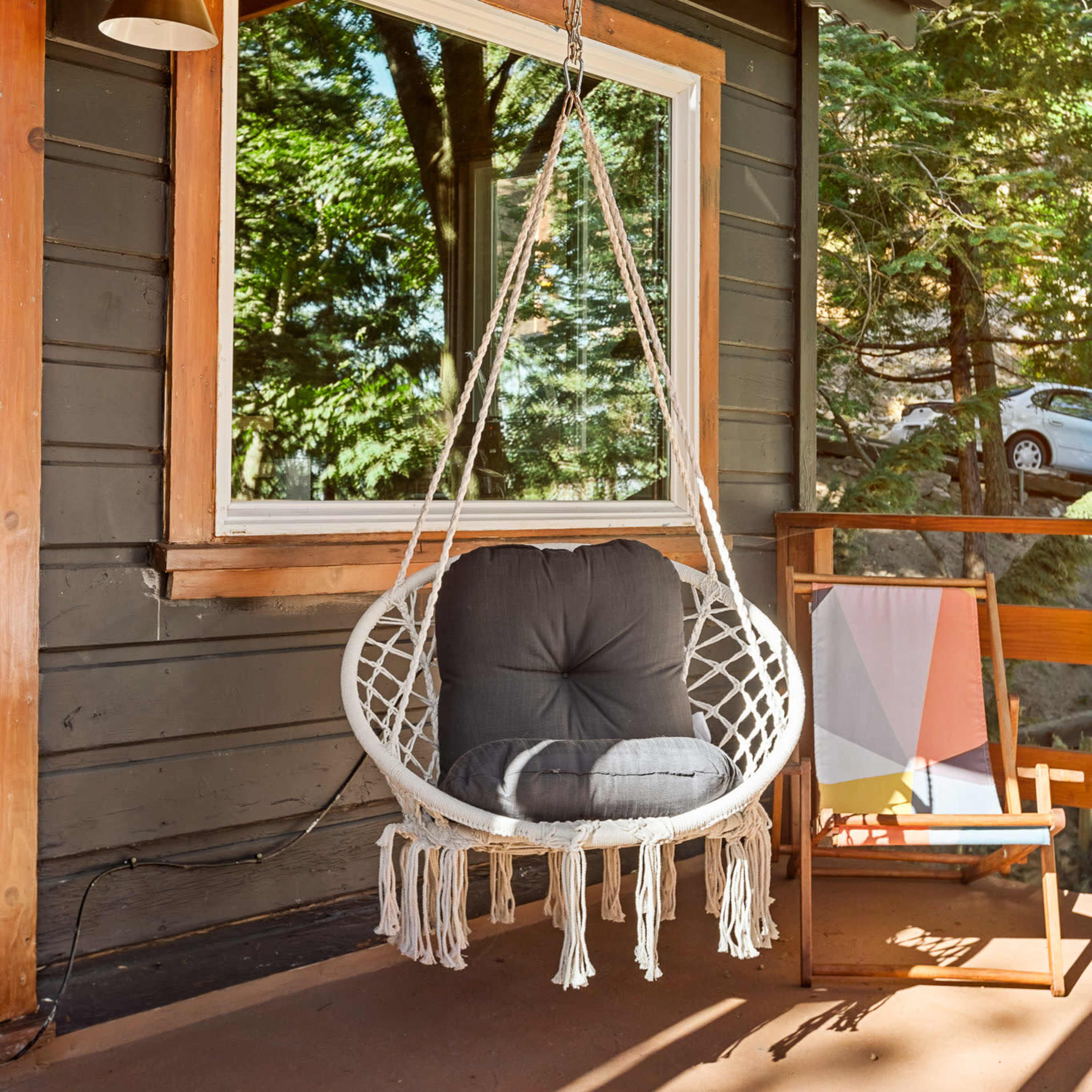 A macramé hanging chair with a cushion is suspended from a wooden porch ceiling next to a window reflecting greenery outside.