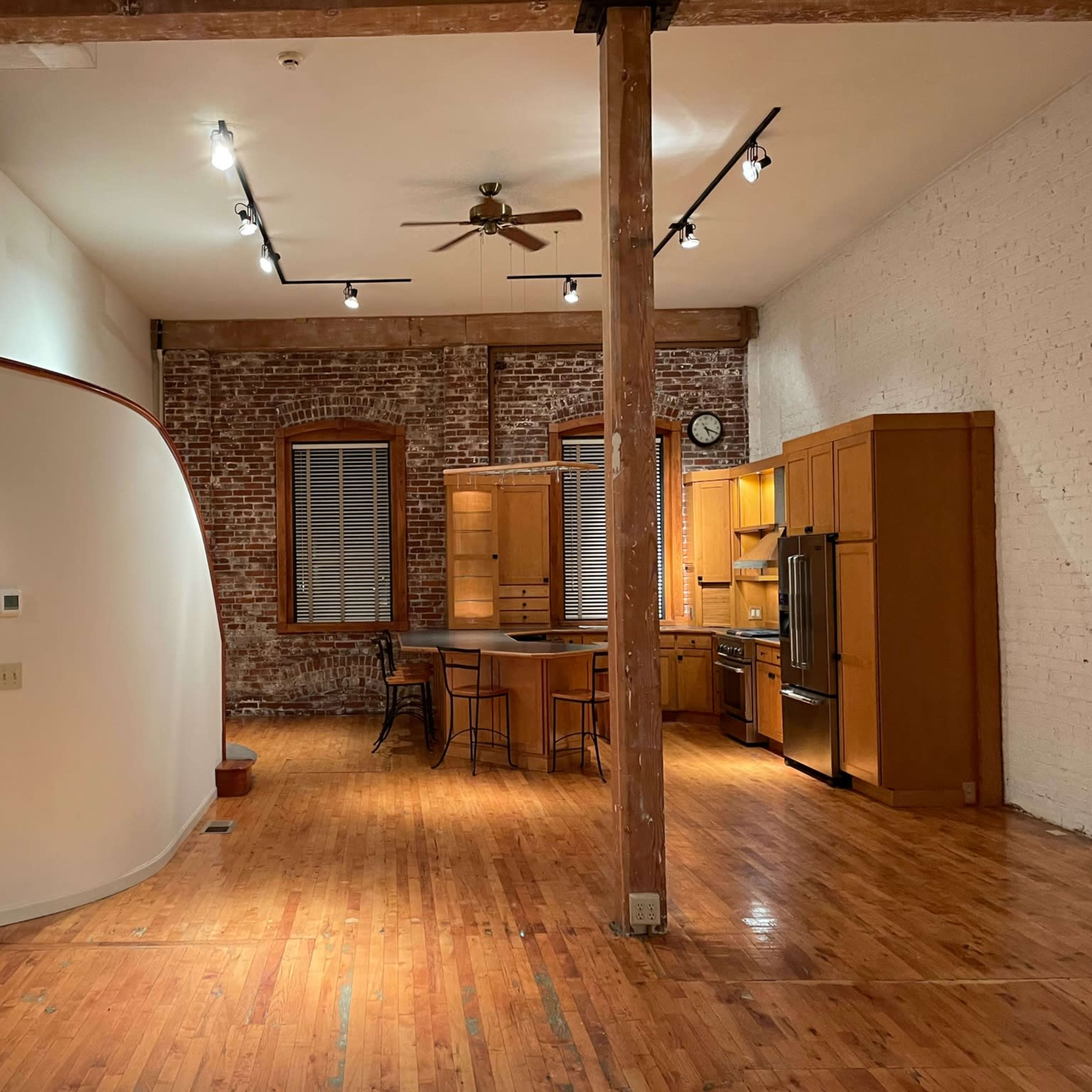 An open living space with wooden flooring, exposed brick walls, and a kitchen featuring wooden cabinets and appliances.