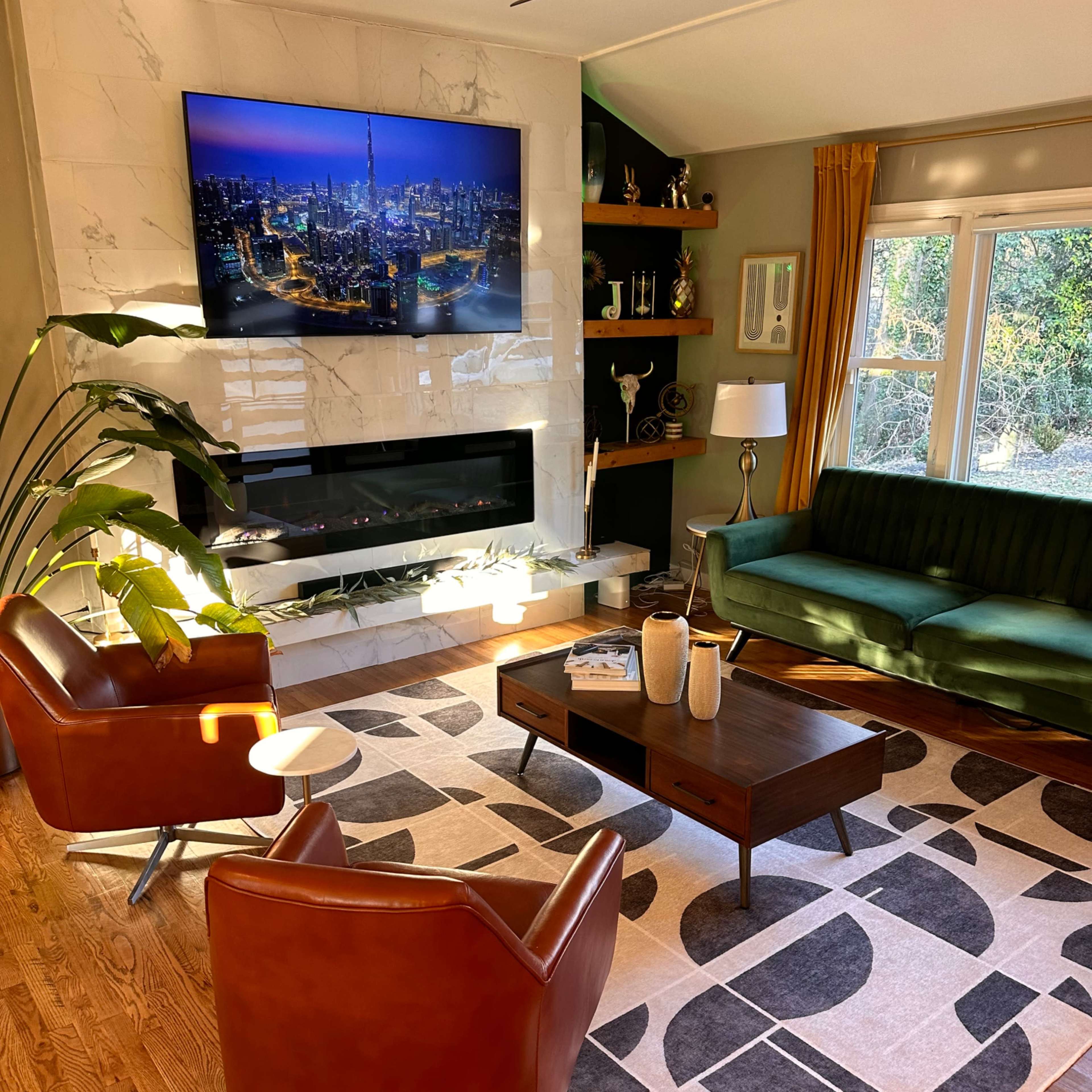 A modern living room featuring a green sofa, two brown armchairs, a wooden coffee table, and a large television mounted above a marble fireplace.