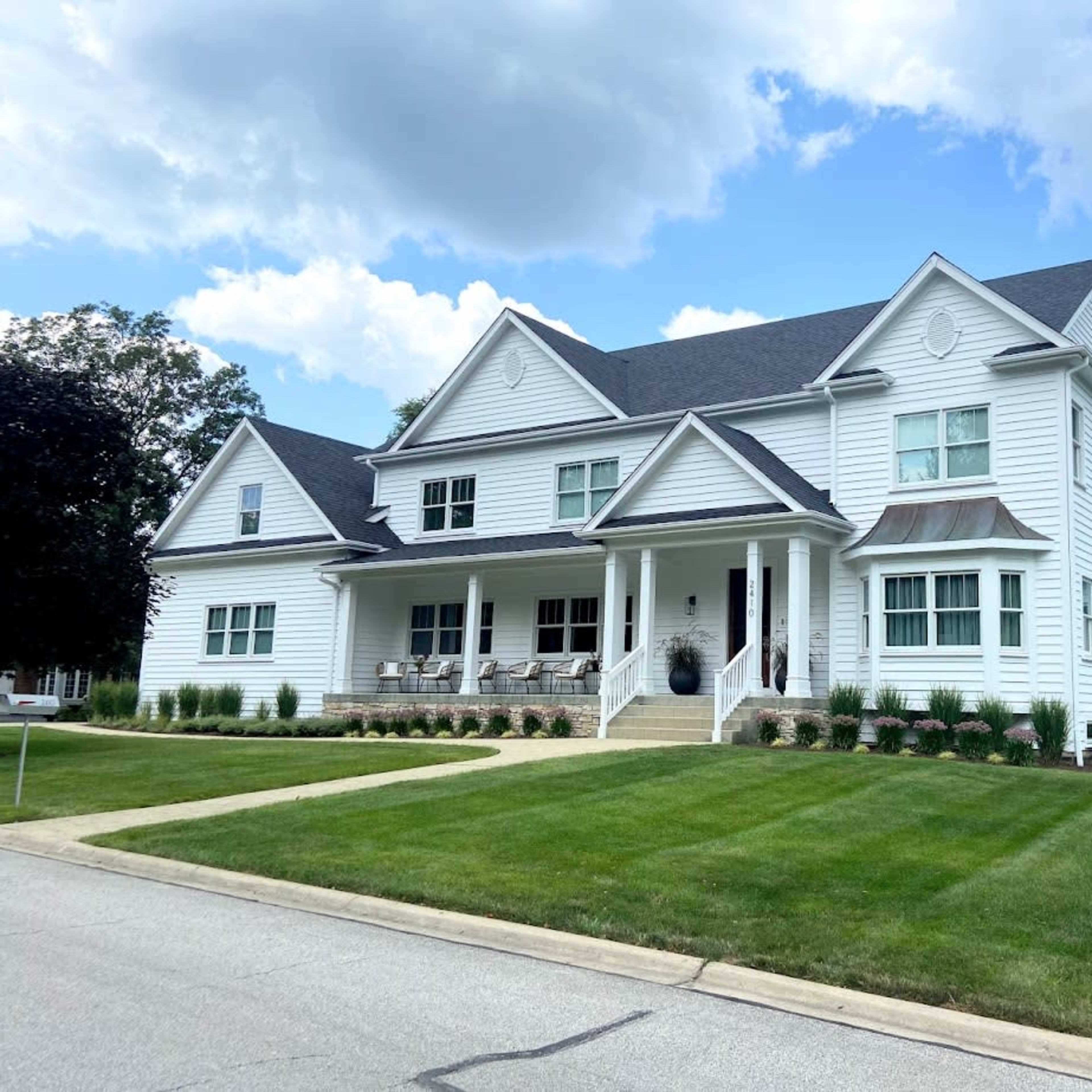 A large, two-story white house with a gray roof, featuring a front porch and well-maintained lawn.