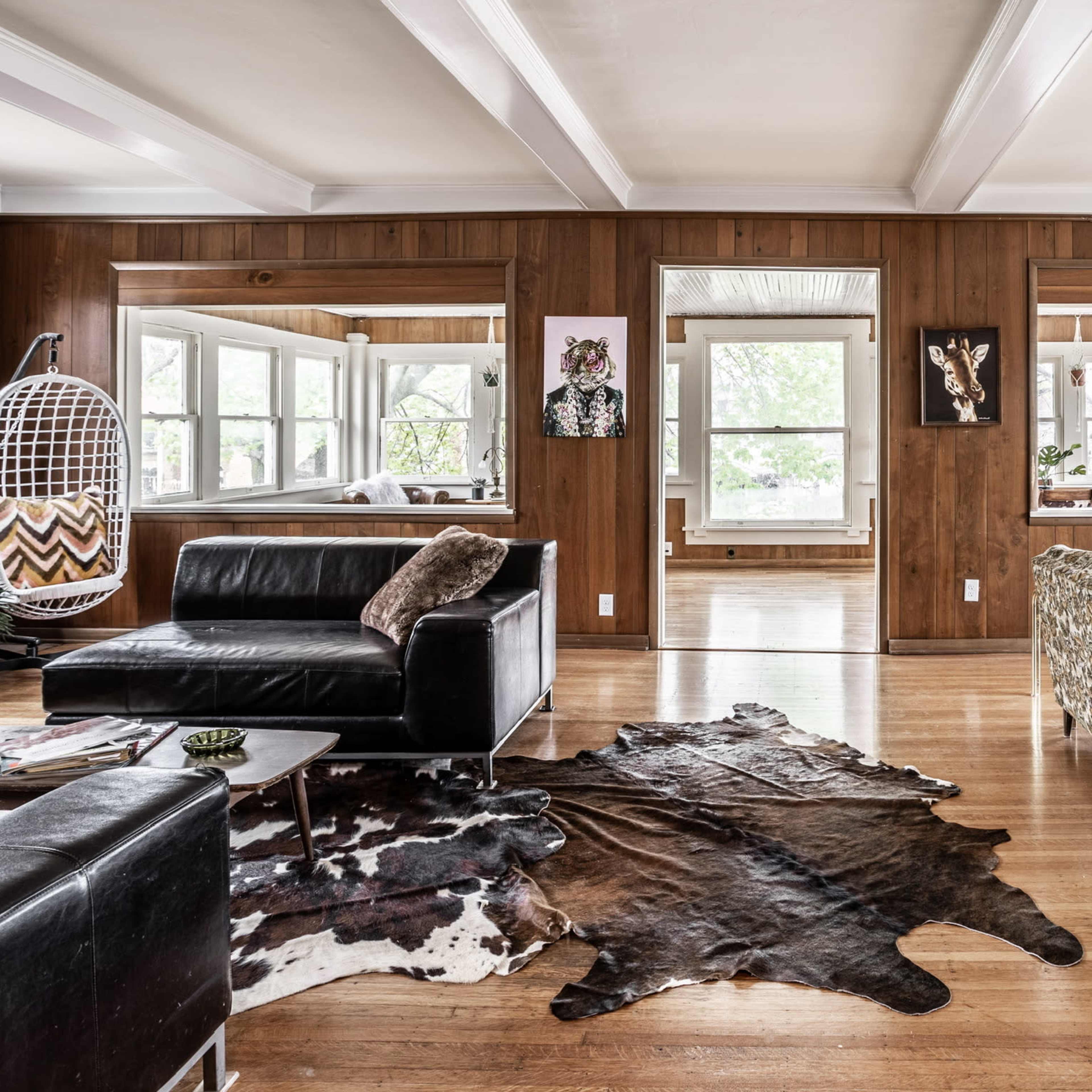 A living room with wooden walls, large windows, and a mix of modern and patterned furniture, including a black sofa and a decorative couch, arranged around a cowhide rug.