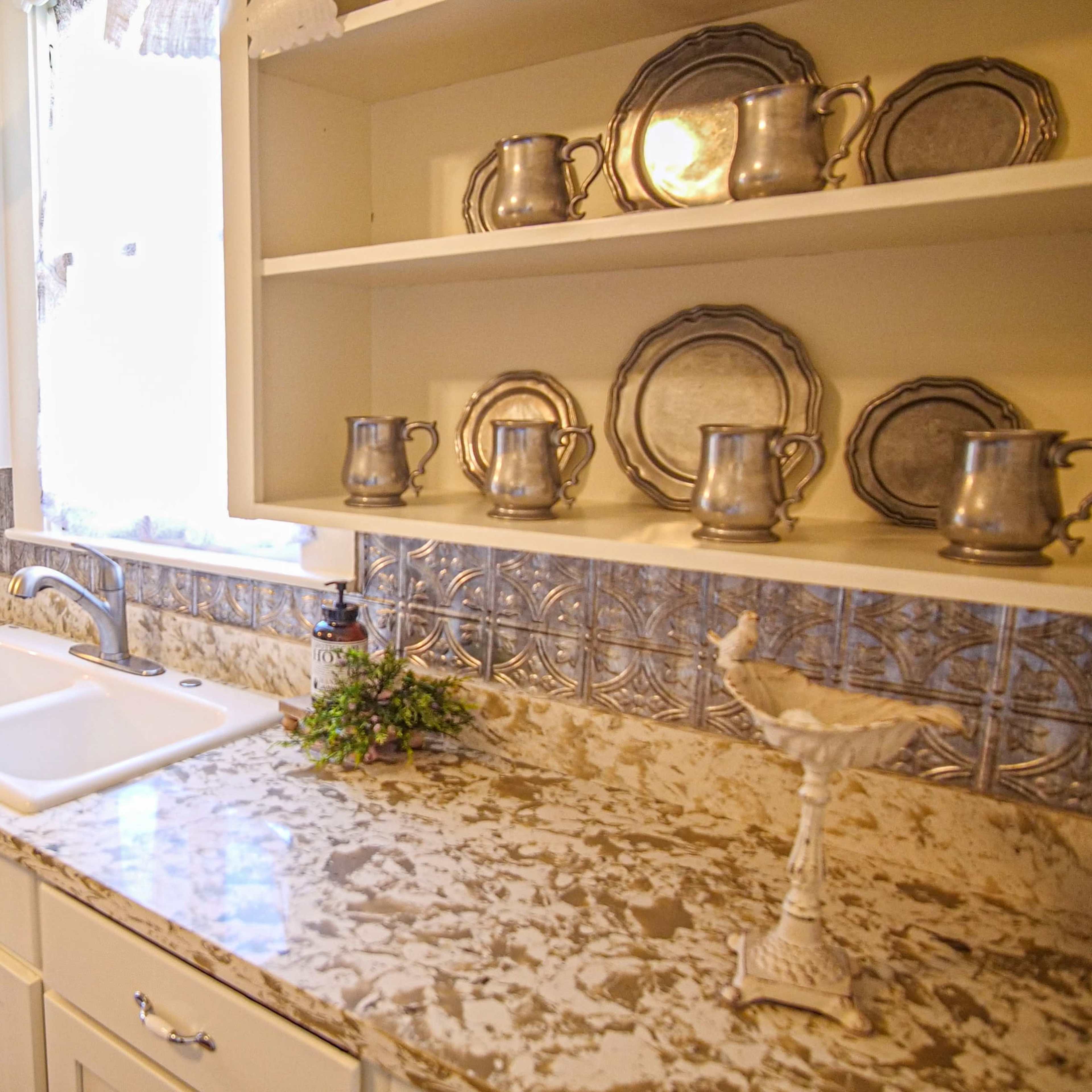 A kitchen with a white sink, marble countertop, open shelves containing silver serving pieces, and a doorway leading to a screened porch.