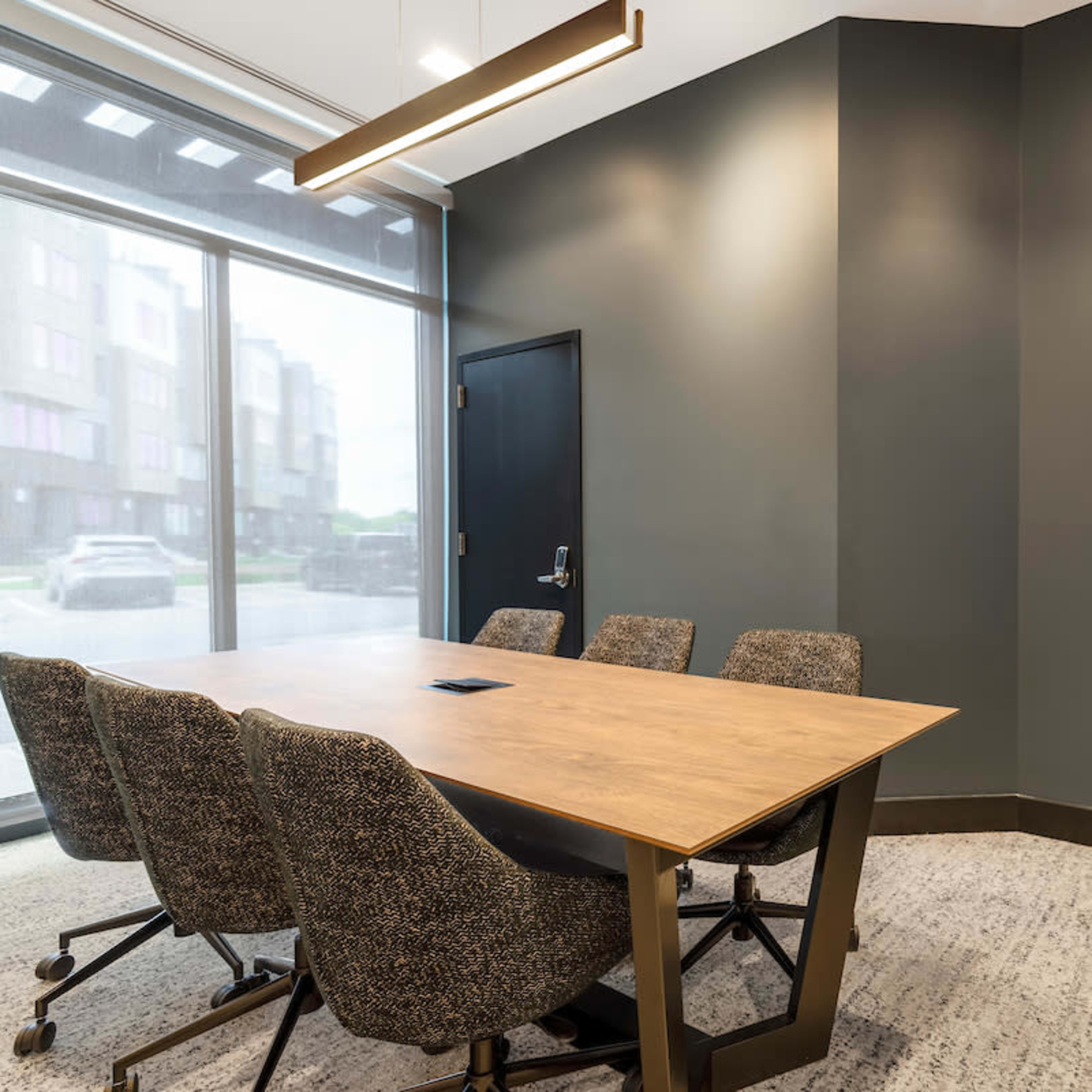 A conference room features a wooden table surrounded by four upholstered chairs and a green moss wall art on the gray wall.