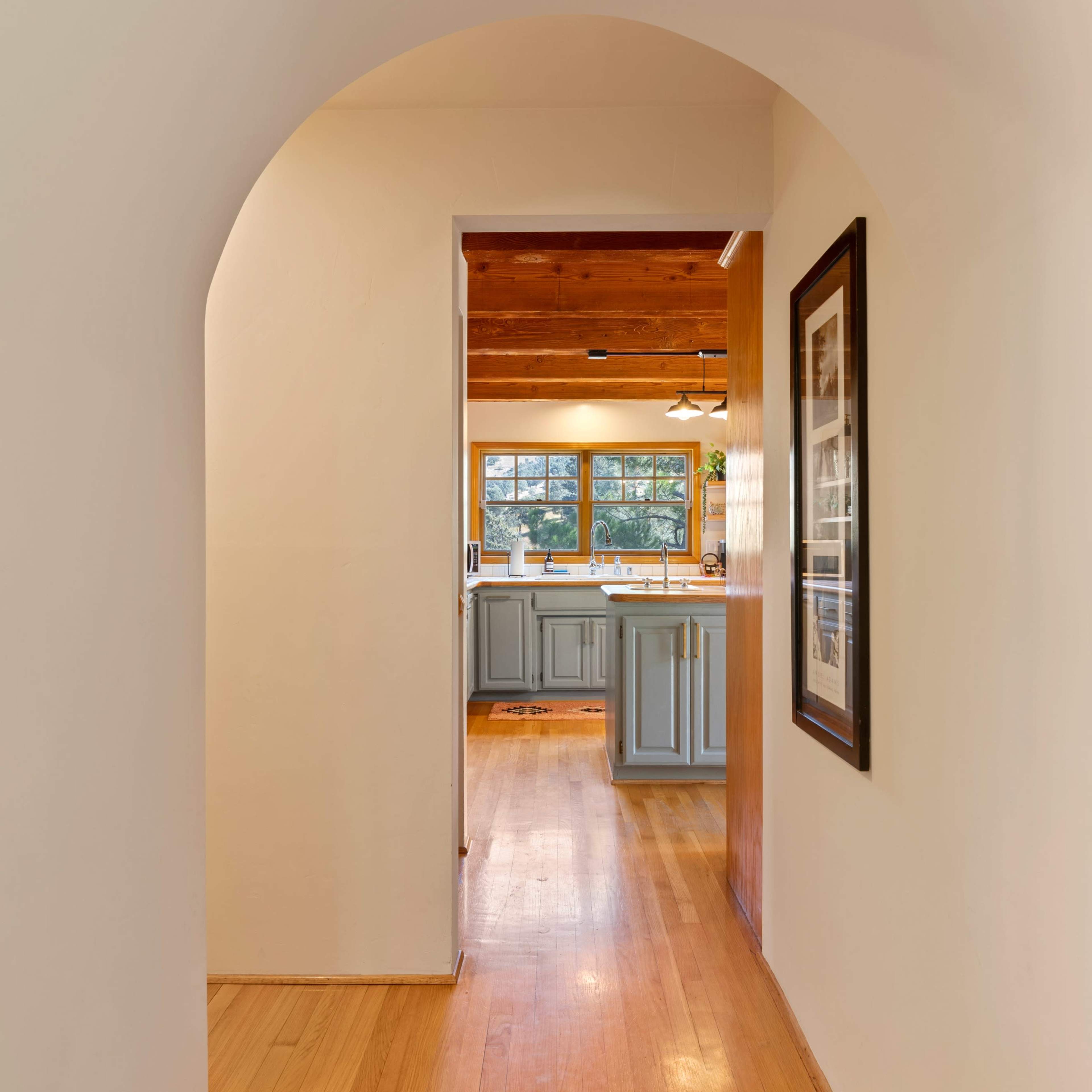 A hallway with a rounded arch leads to a kitchen with wooden beams and light-colored cabinetry.
