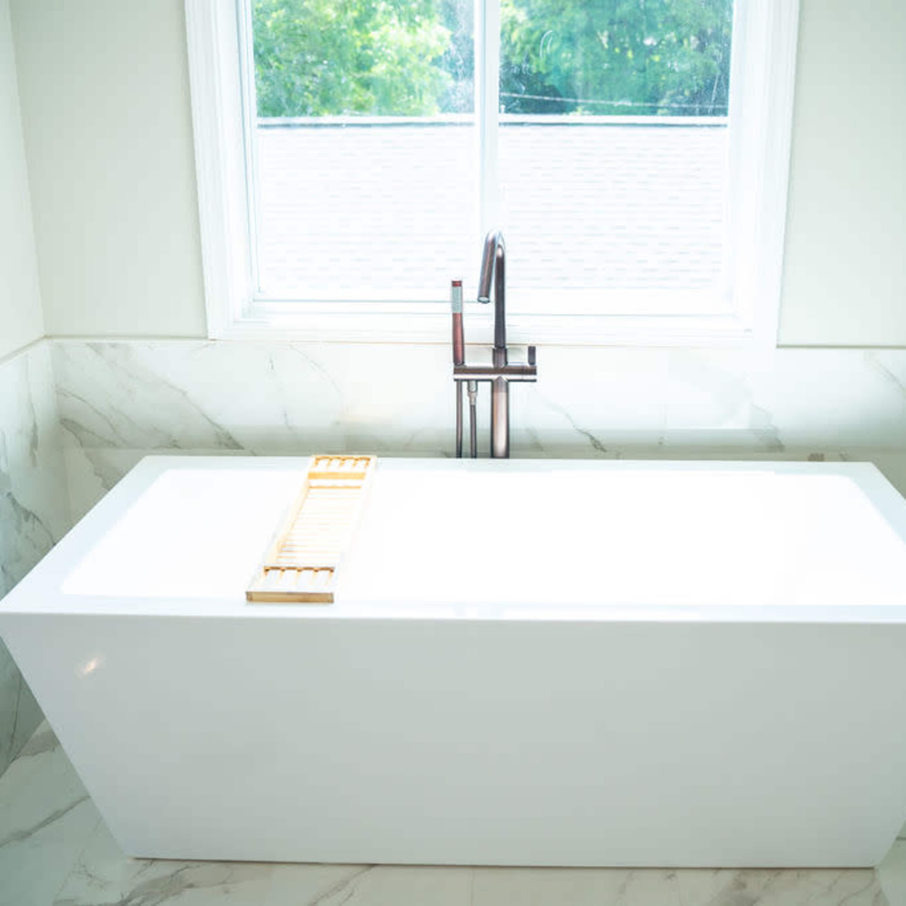 A modern white freestanding bathtub is positioned next to a window, with a wooden bath tray resting on its rim.