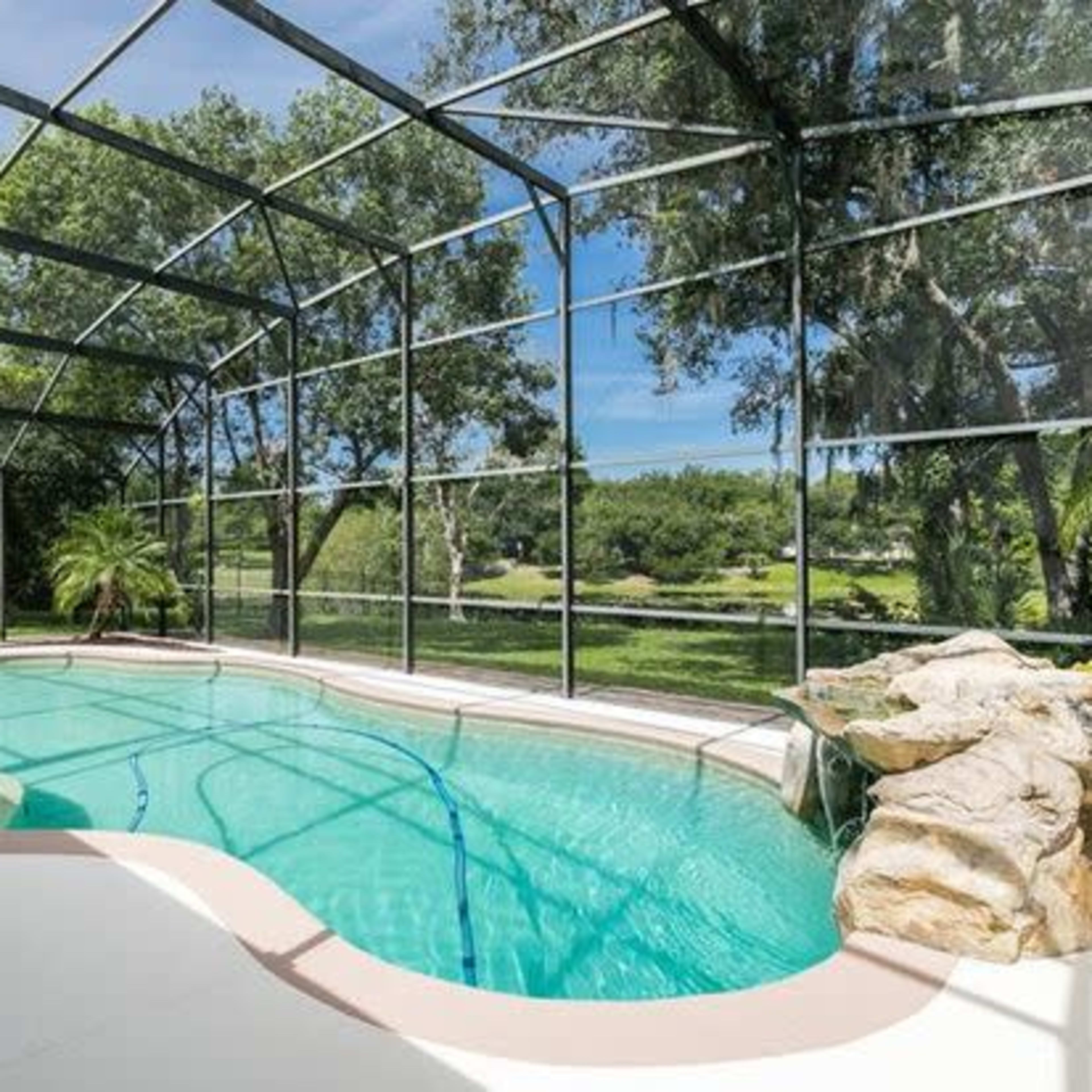 A screened-in pool area with a curved, turquoise pool surrounded by greenery and a rock feature.