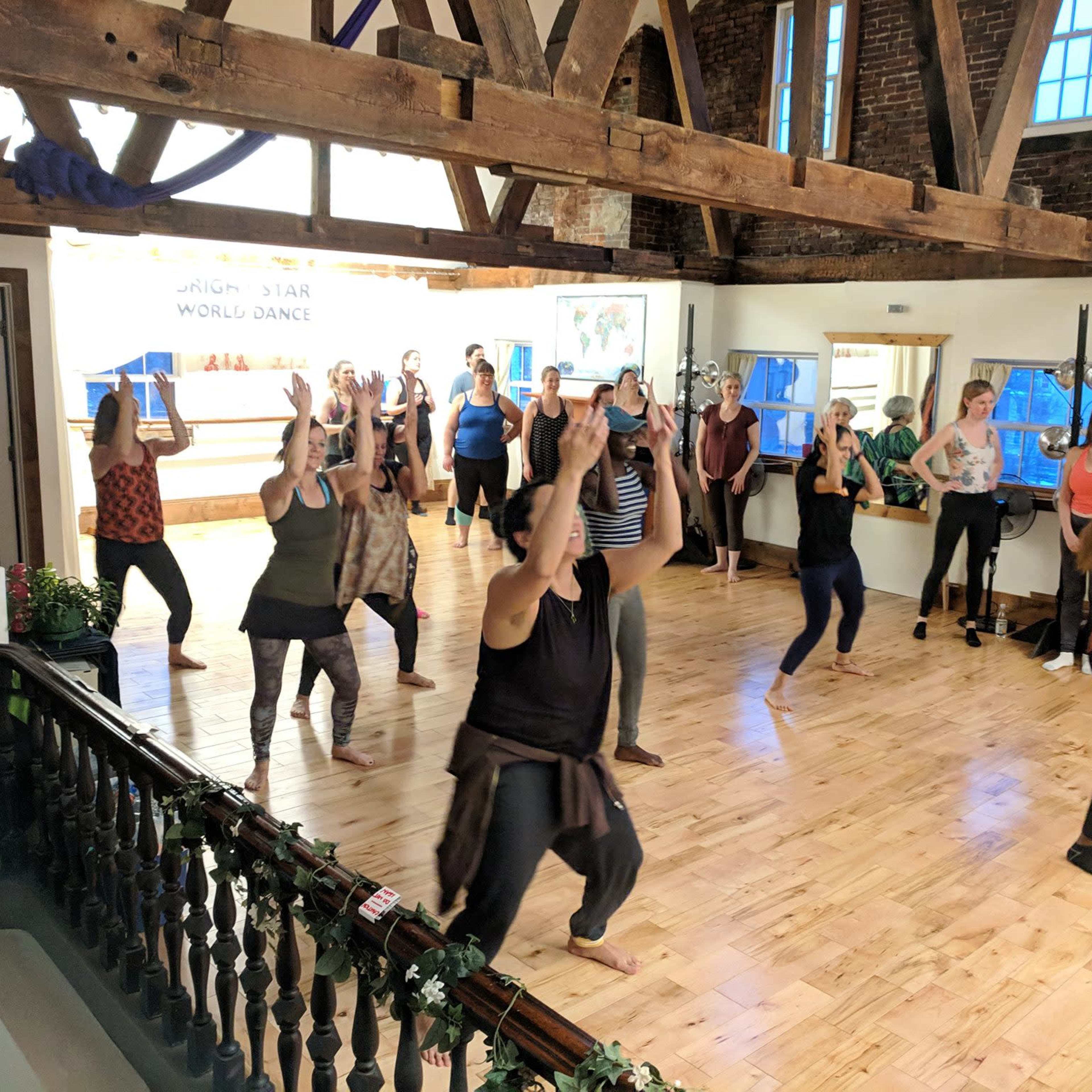 A group of people participate in a dance class in a spacious studio with wooden beams and large windows.