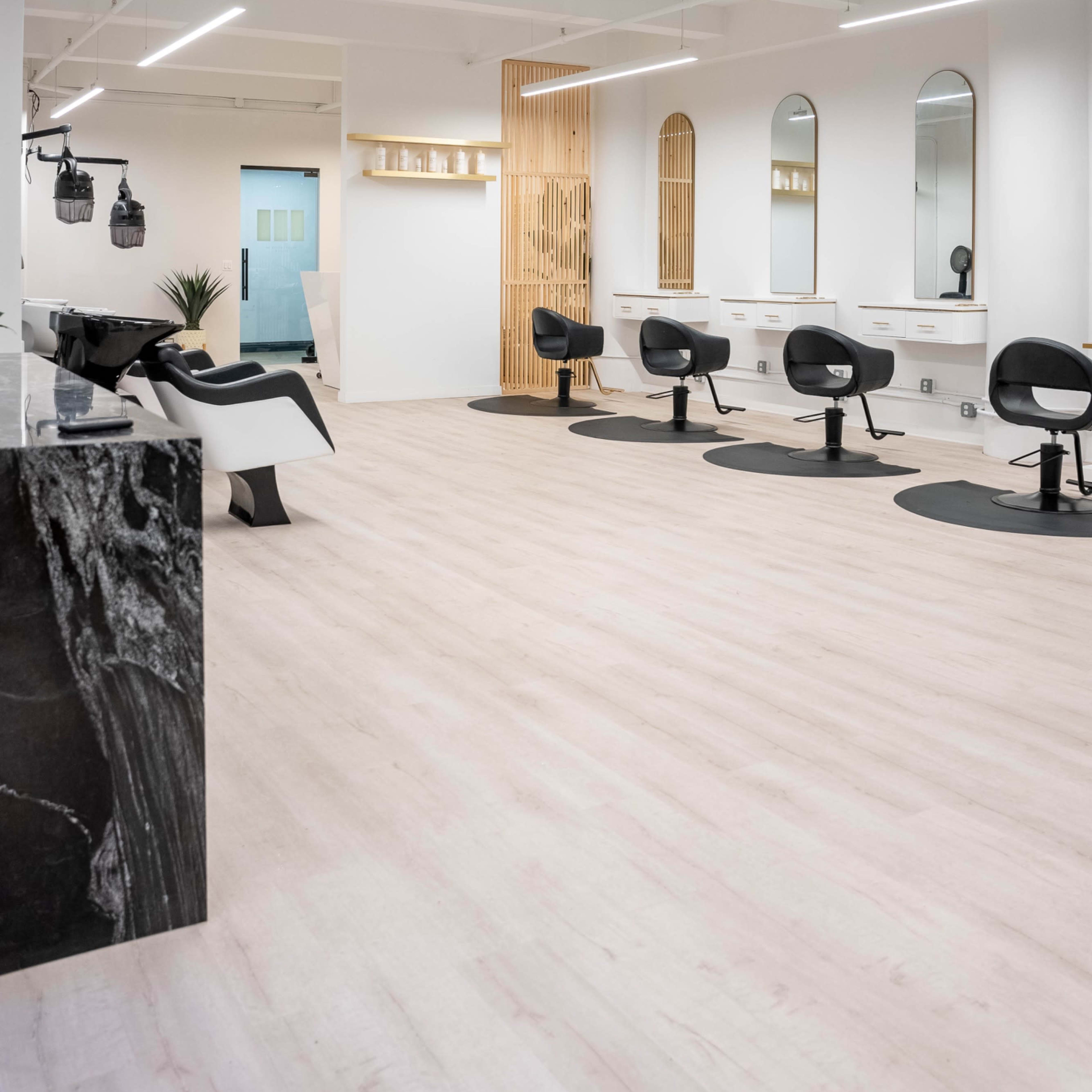 A modern hair salon with several styling stations, mirrors, and black salon chairs arranged on light-colored wooden flooring.