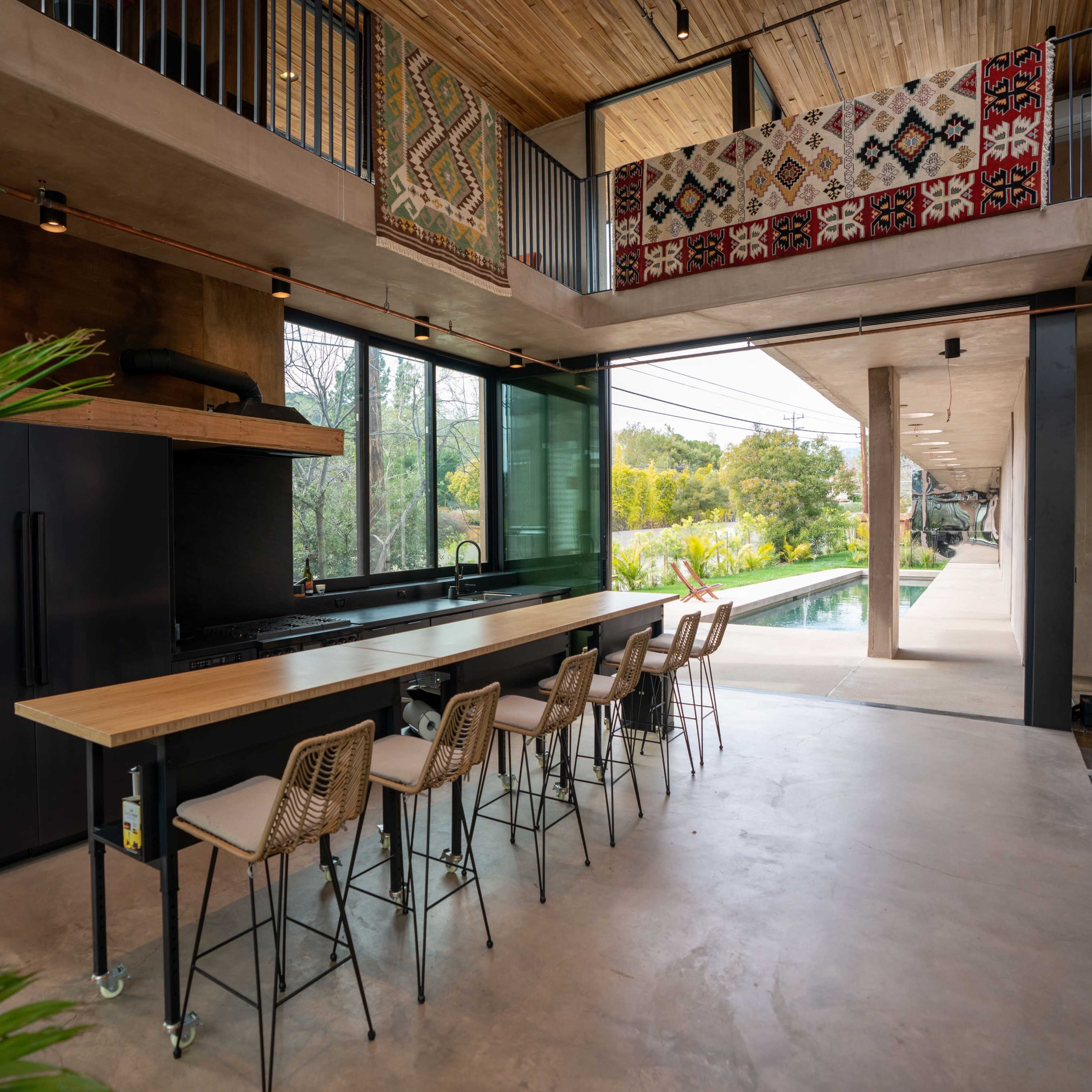 A modern kitchen with a large wooden countertop, several high stools, and large windows overlooking a green outdoor space.