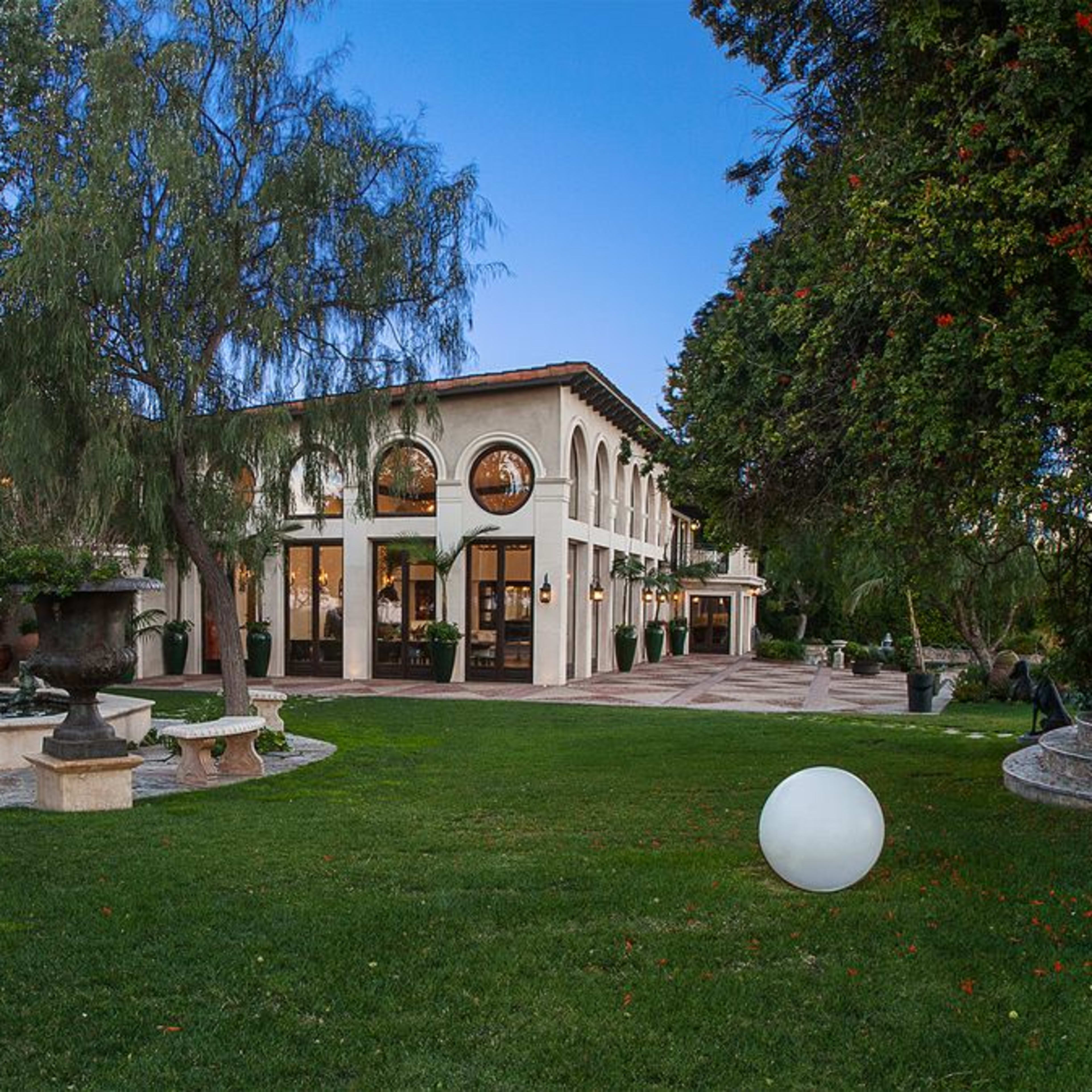A large, two-story house with arched windows surrounded by a green lawn and trees, featuring a pool and decorative stone landscaping.