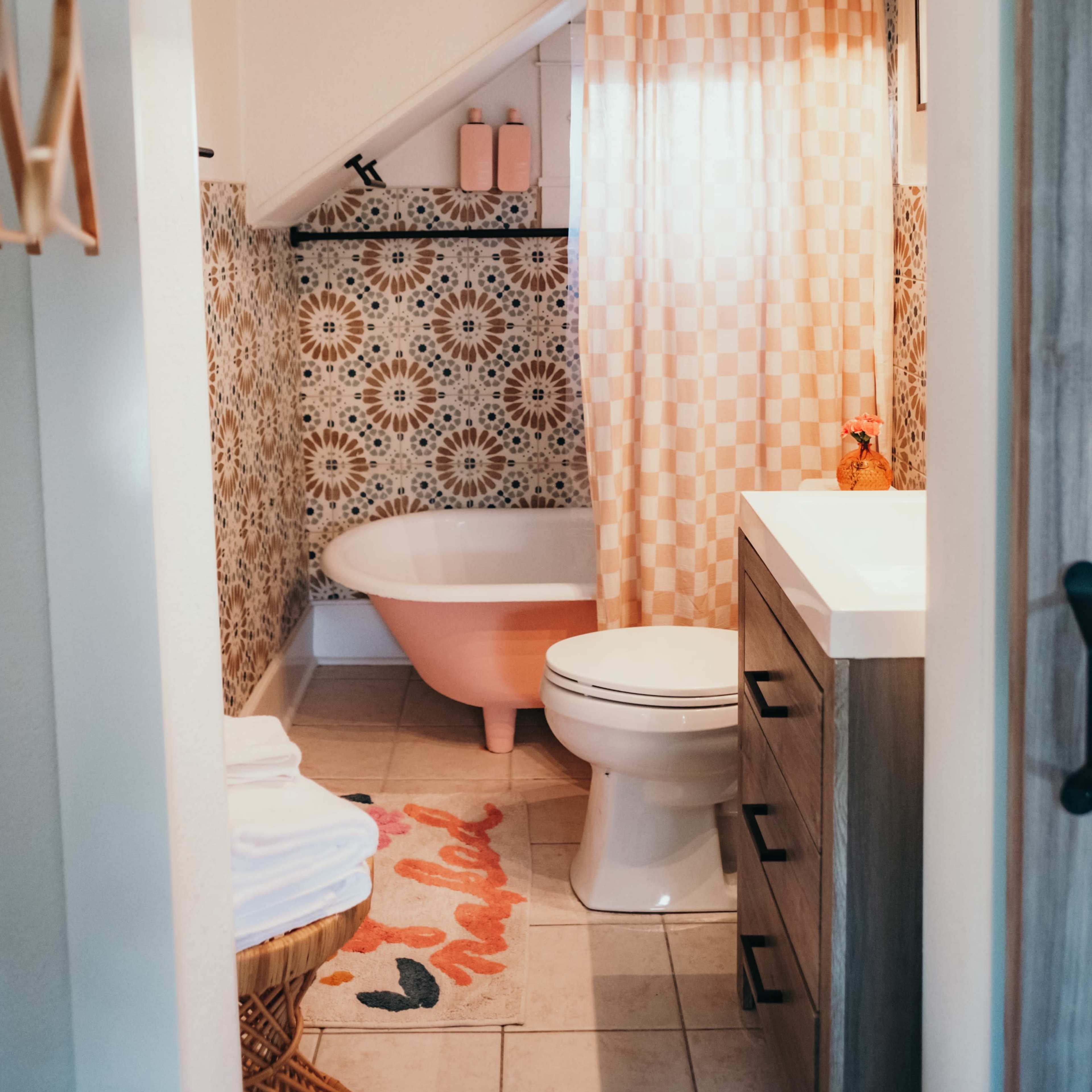 A small bathroom featuring a pink bathtub, a white toilet, a sink with a wooden cabinet, and patterned wallpaper under a sloped ceiling.
