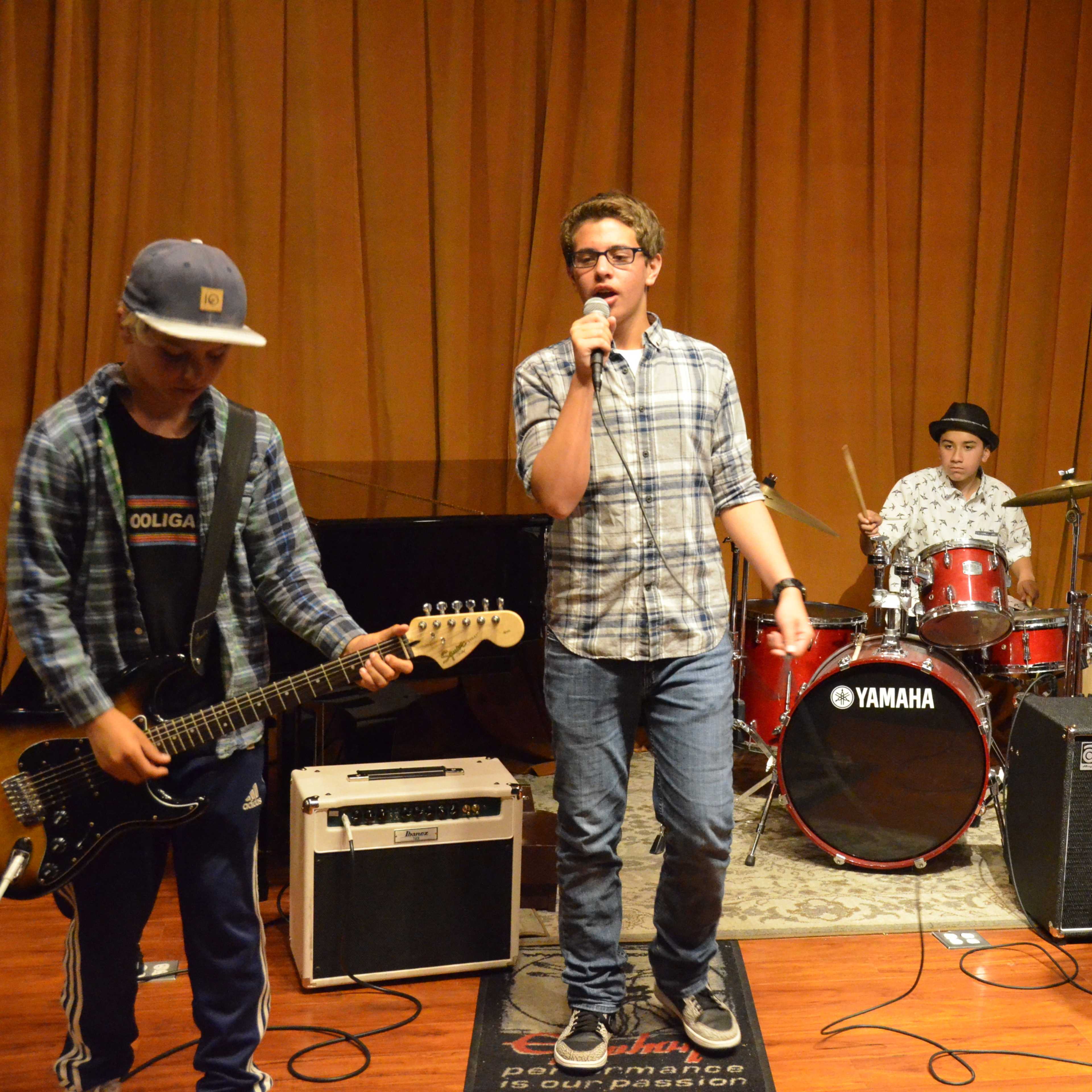 A group of four musicians performs in a studio, with one singer holding a microphone while the others play guitar, bass, and drums.