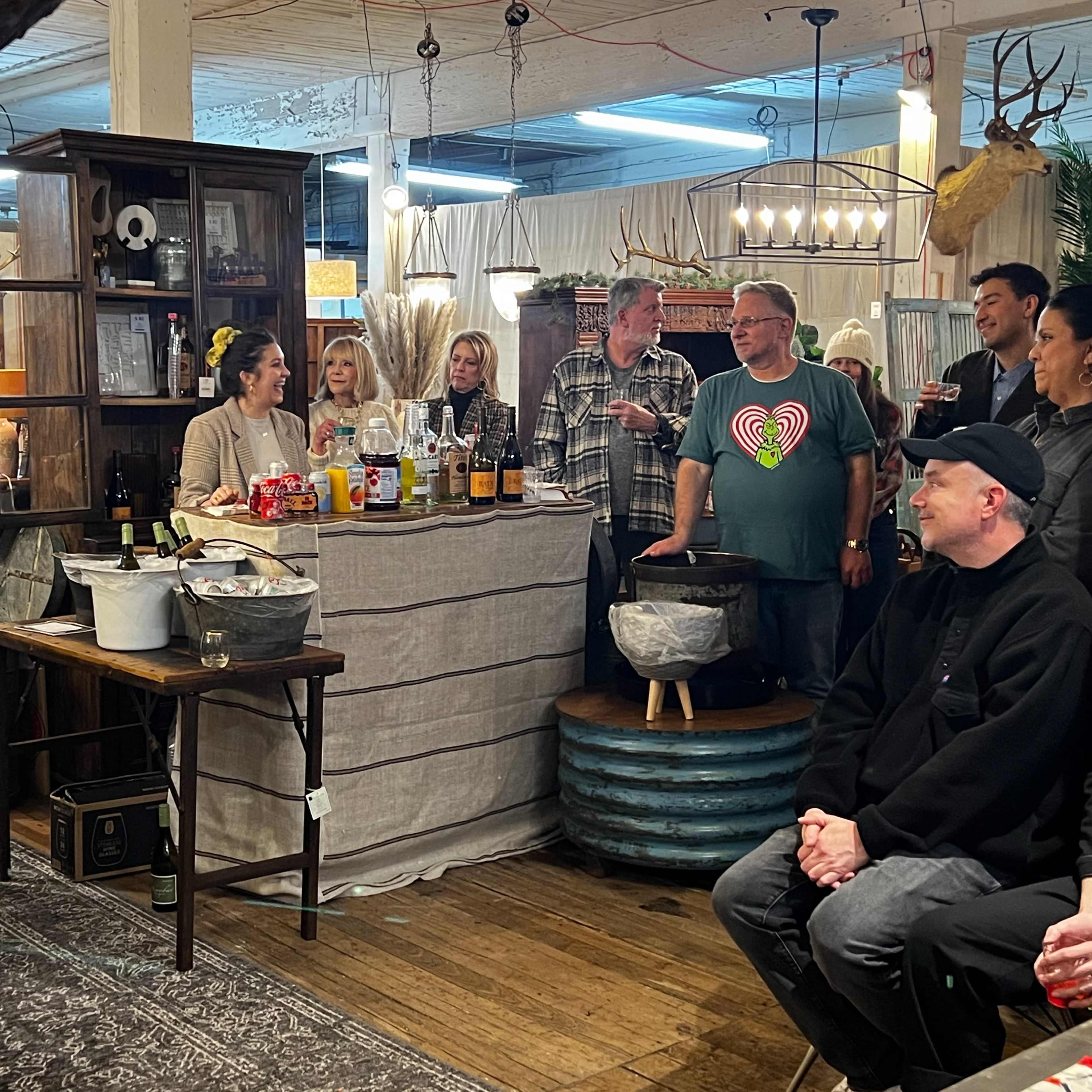 A group of people gathers around a table in a decorated store while a woman speaks, with various food and drink items displayed on the table.
