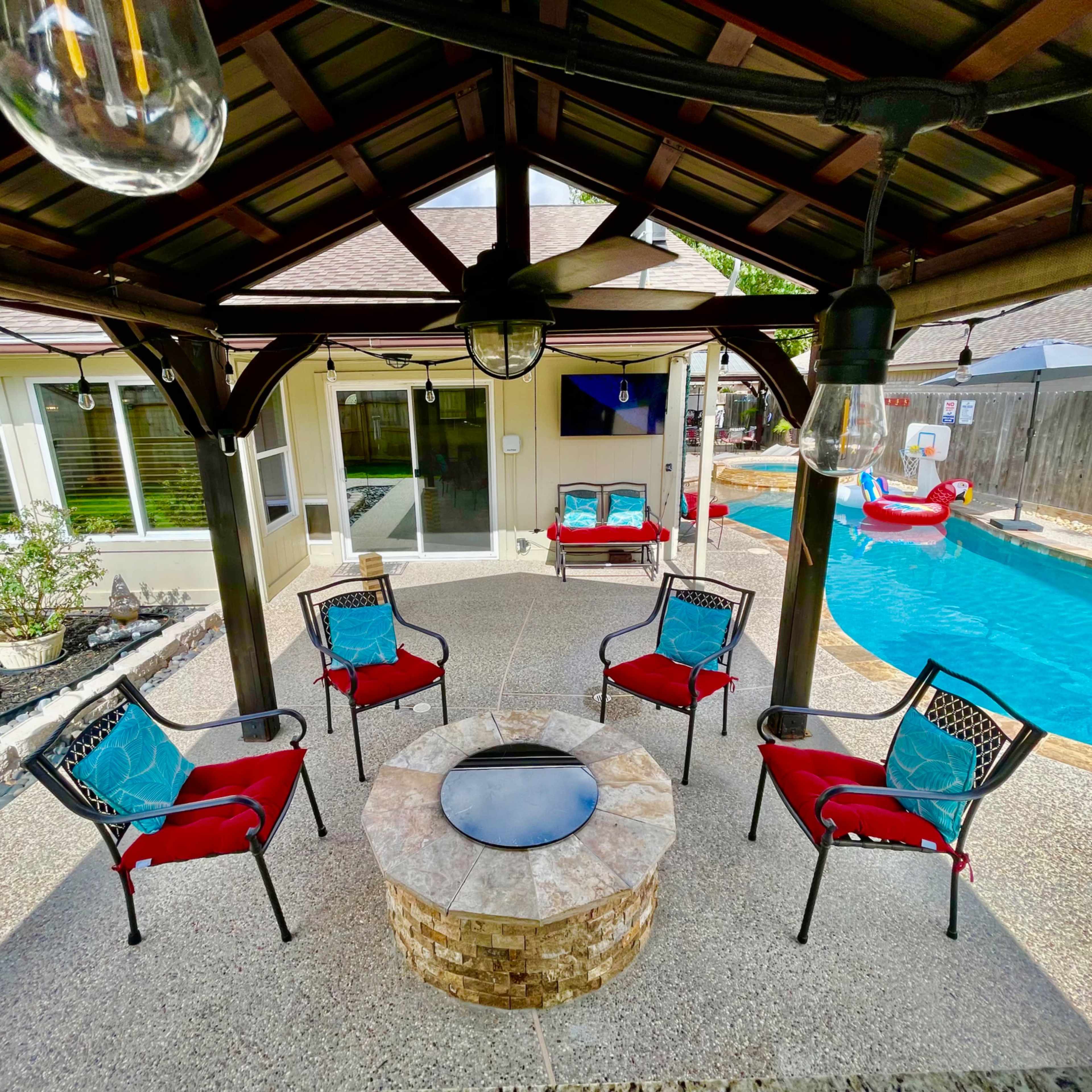 A covered patio features six red chairs arranged around a stone fire pit, with a swimming pool visible in the background.