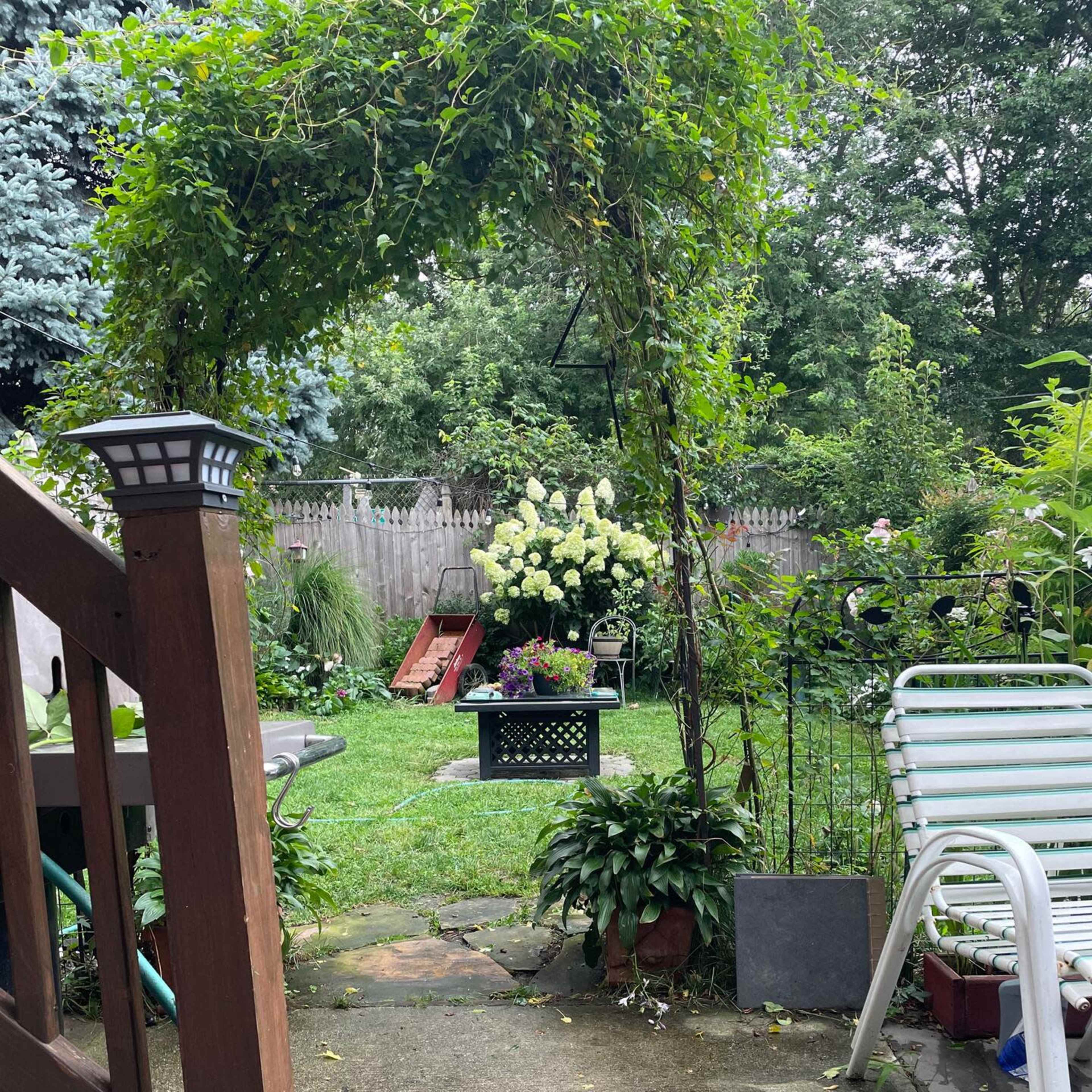A stone path leads from a wooden deck into a green backyard featuring various plants, a planter with white flowers, and a metal chair.