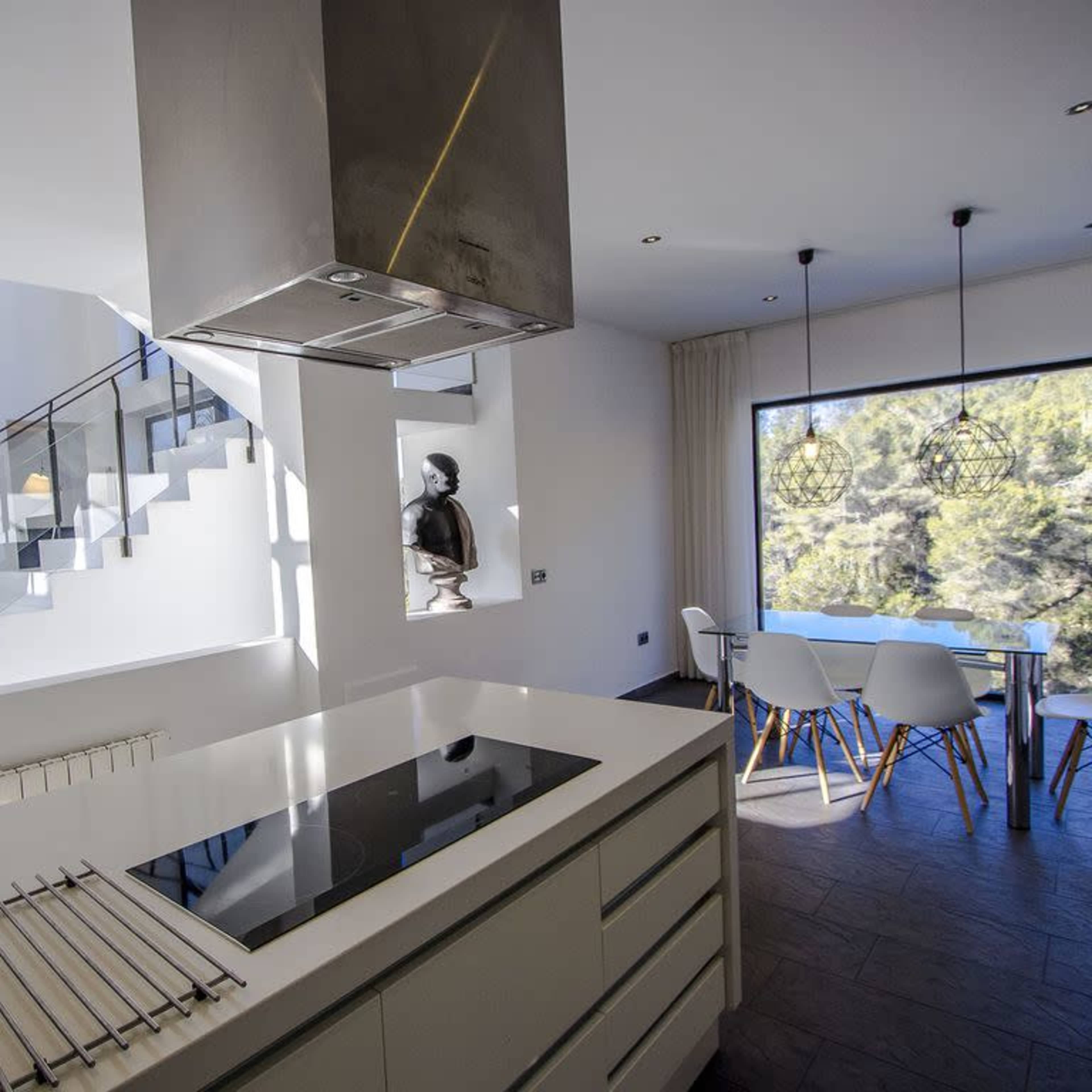 A modern kitchen features a large island with a stovetop, industrial-style hood, and a dining area with chairs beside a large window.