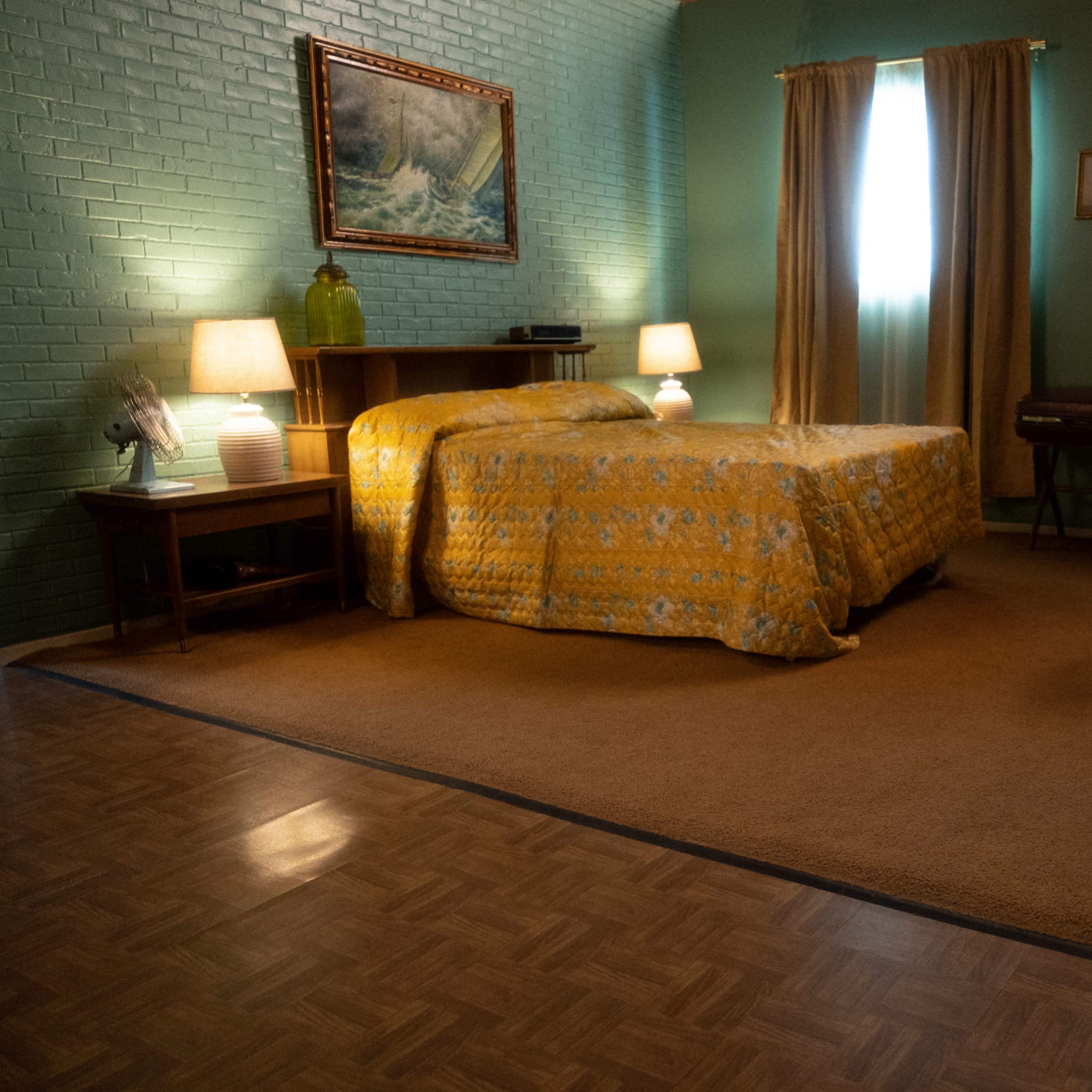 A simple bedroom with a yellow patterned bedspread, a wooden desk, and two lamps beside the bed, illuminated by natural light from a window.