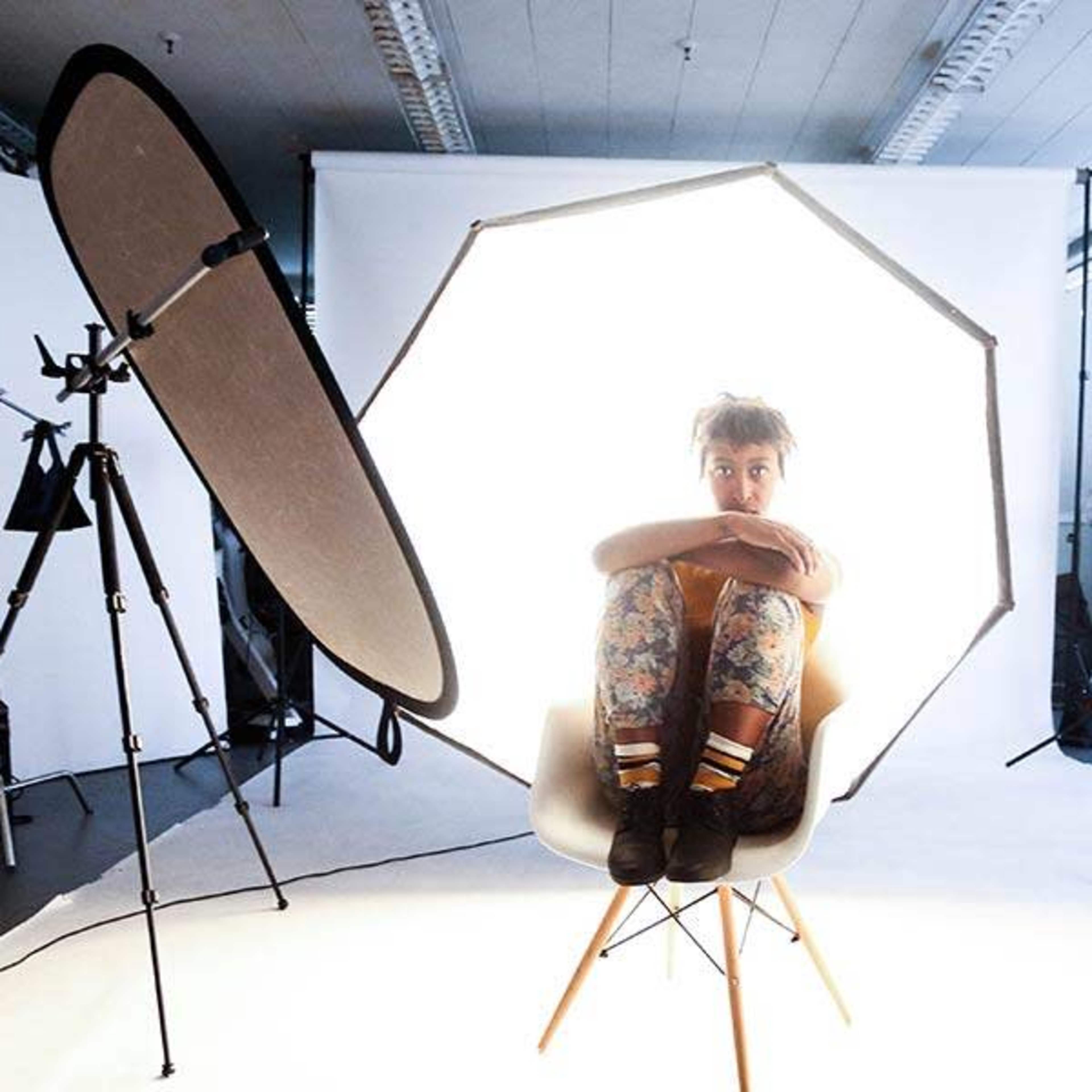 A person sits cross-legged in a chair, surrounded by bright light from a large softbox in a photography studio.