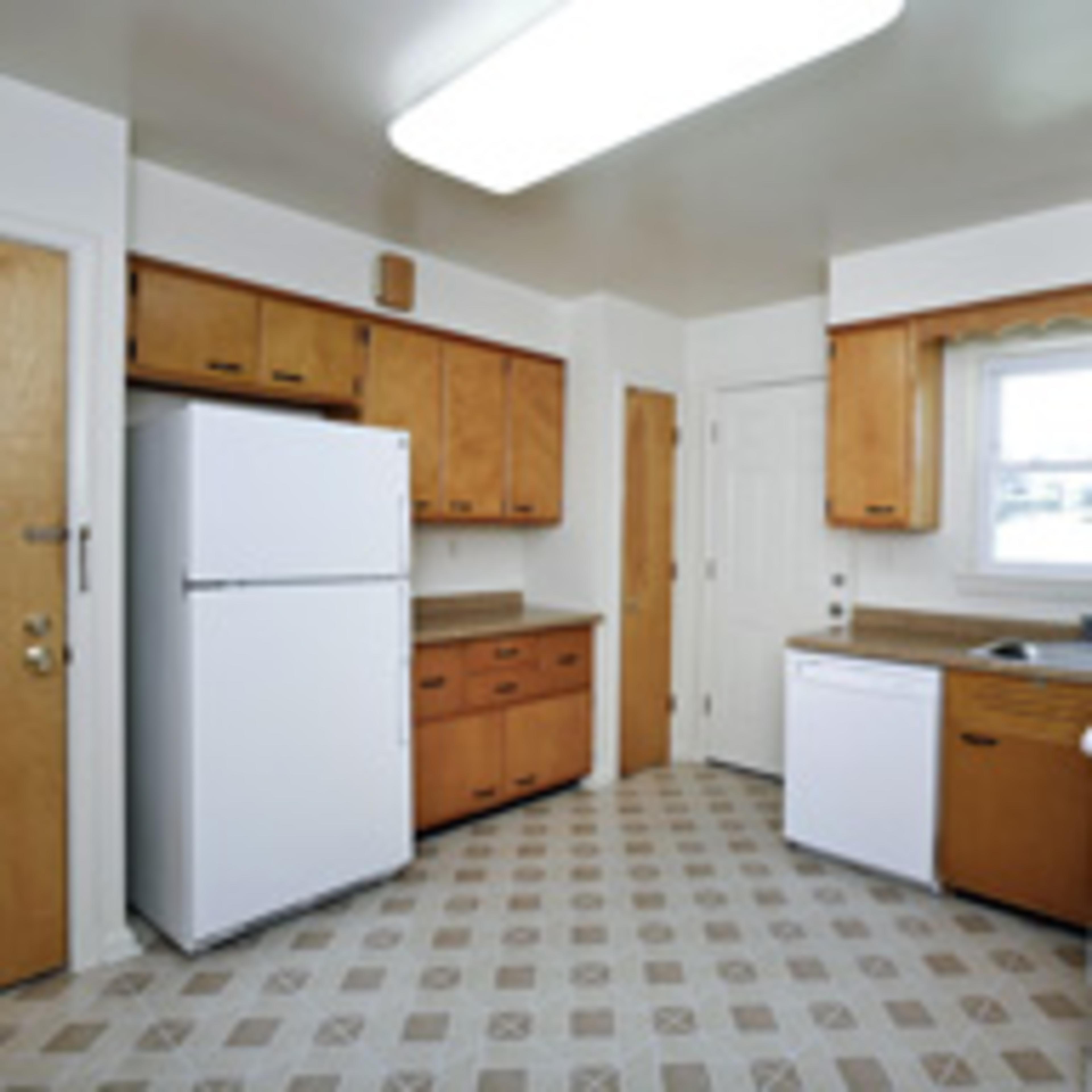 A compact kitchen featuring wooden cabinets, a white refrigerator, a dishwasher, and an oven with a patterned floor.
