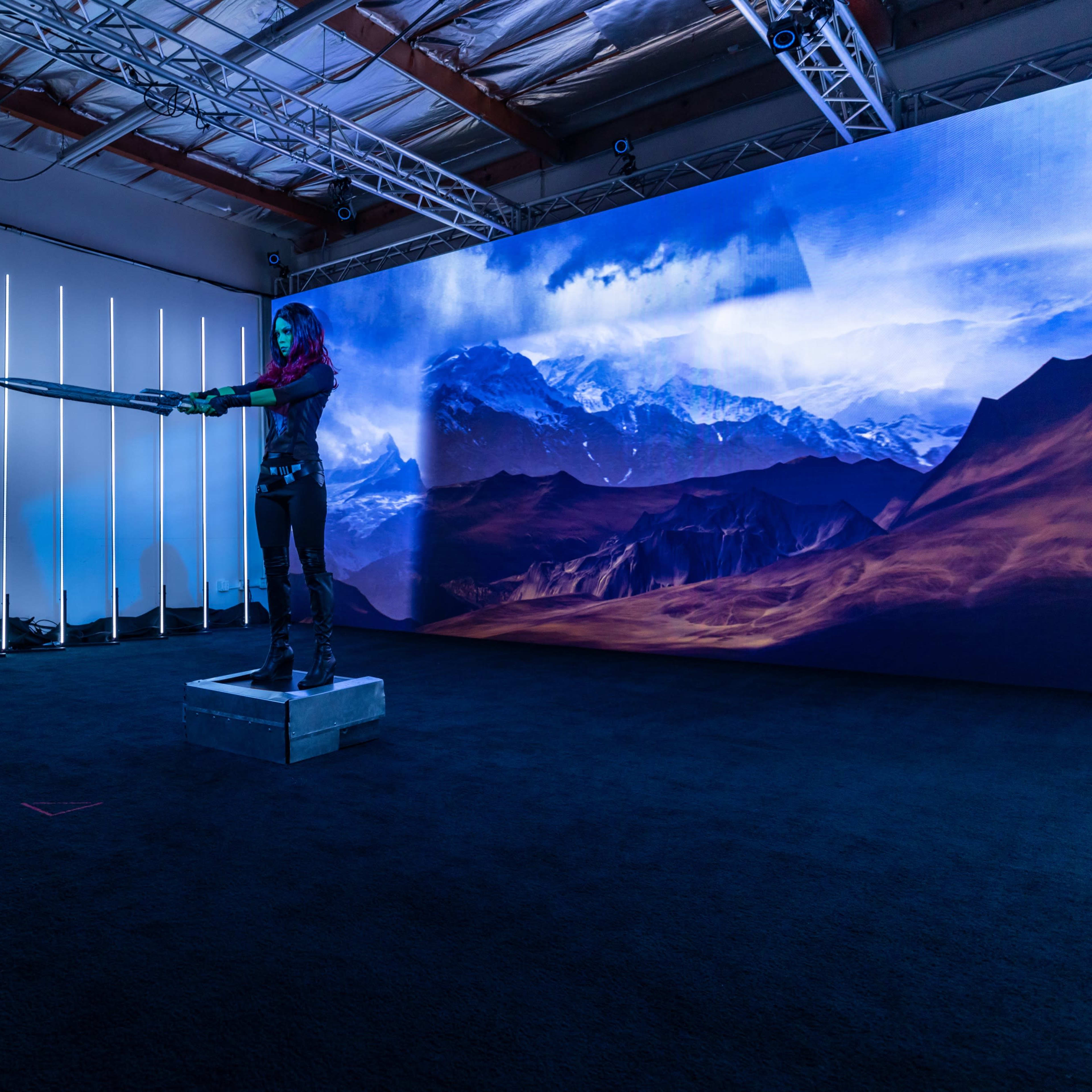A person stands on a platform holding a sword in a studio set designed with a large mountain backdrop and studio lighting.