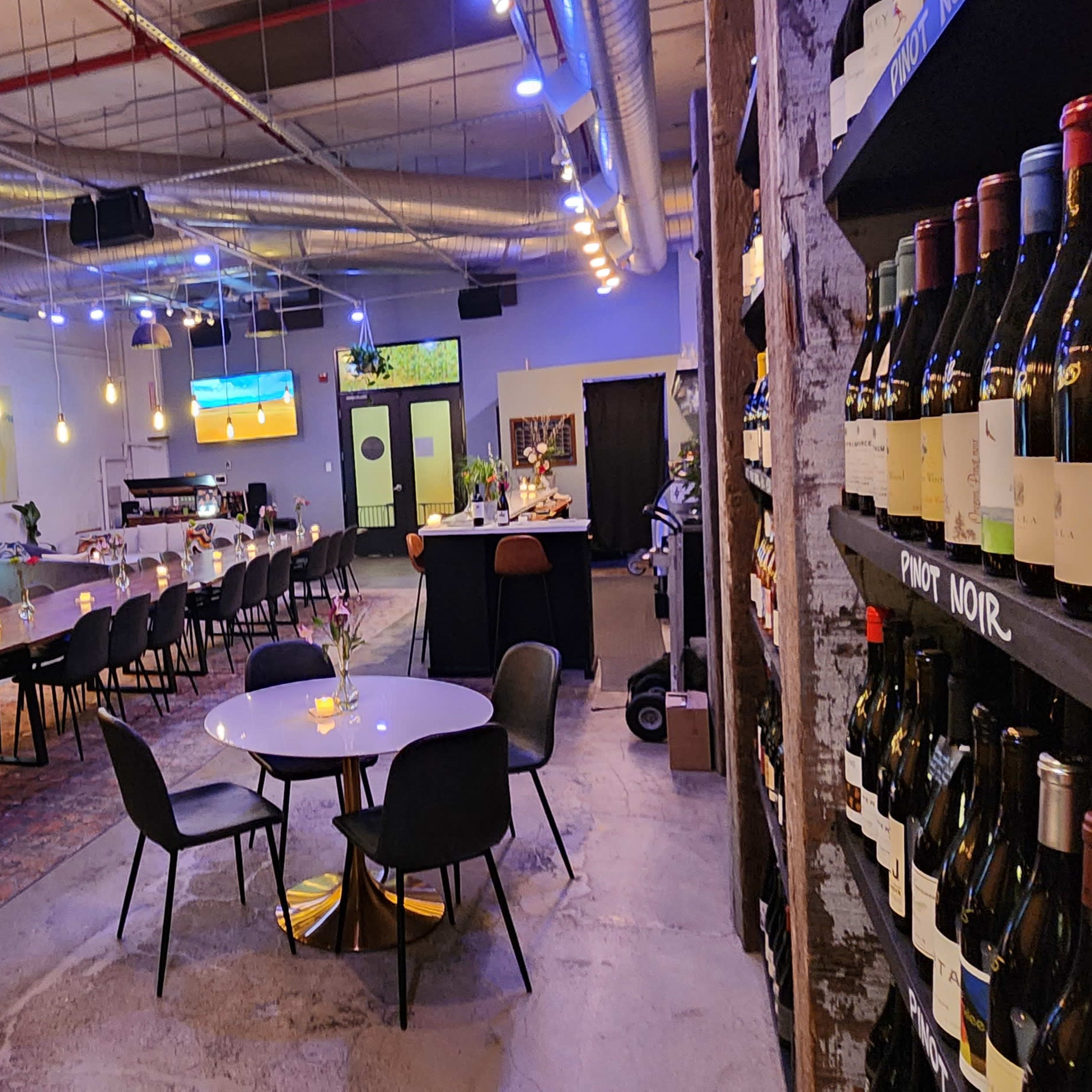 The interior of a modern wine bar featuring a long communal table, several smaller tables, a wine rack displaying numerous bottles, and a bar area in the background.