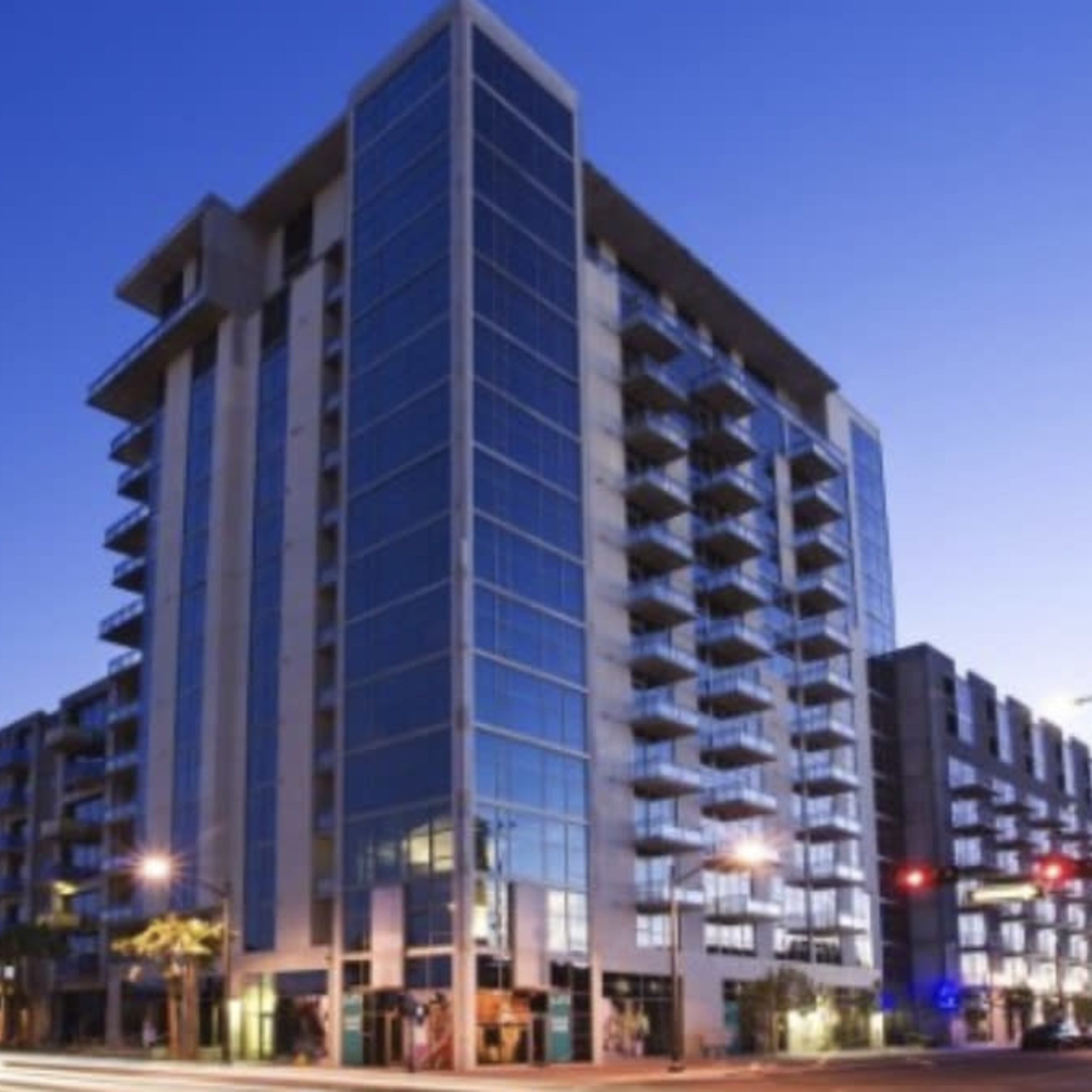 A modern multi-story apartment building with large glass windows is located at a busy intersection during twilight.
