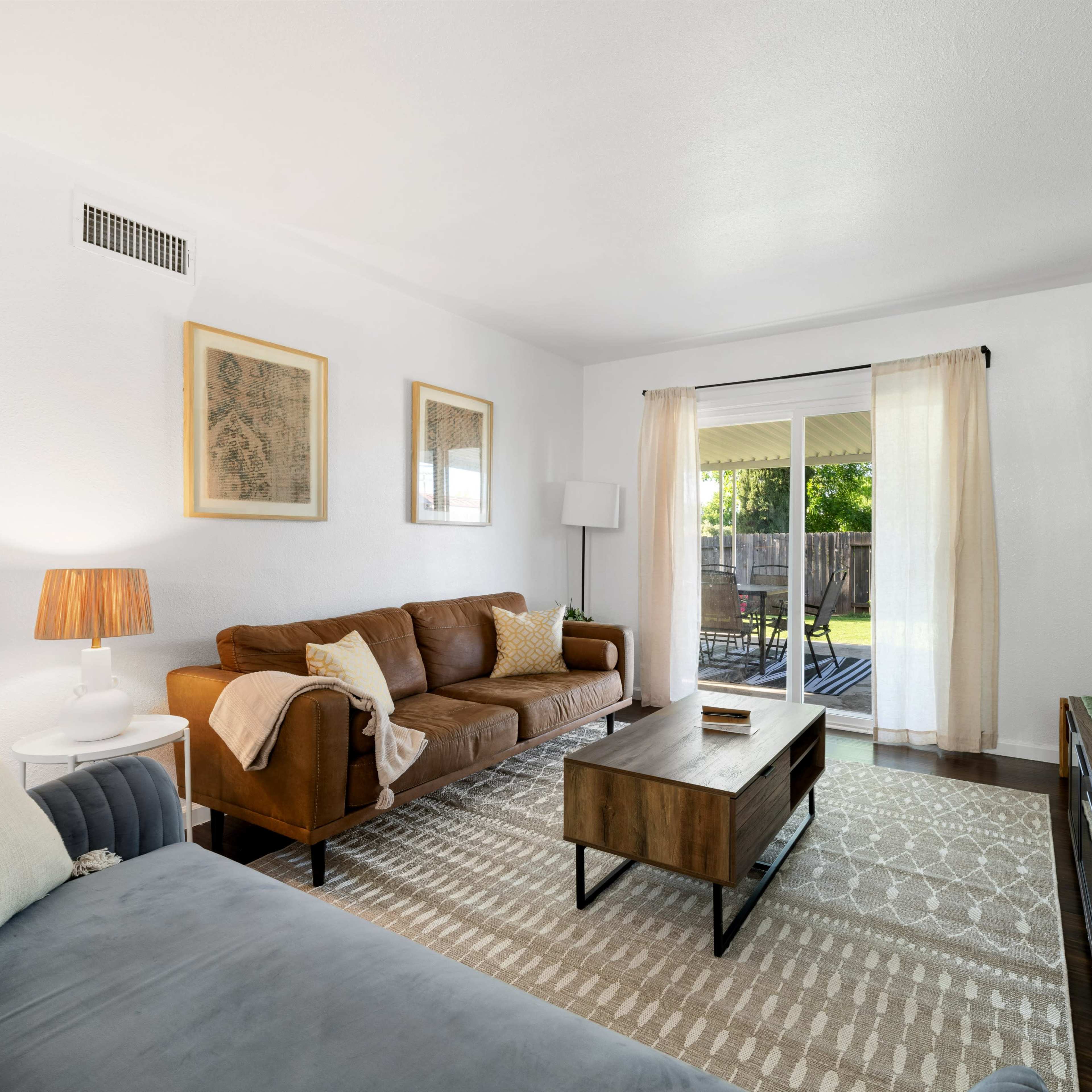 A bright living room with two sofas, a coffee table, a television, and large windows leading to a patio in the background.