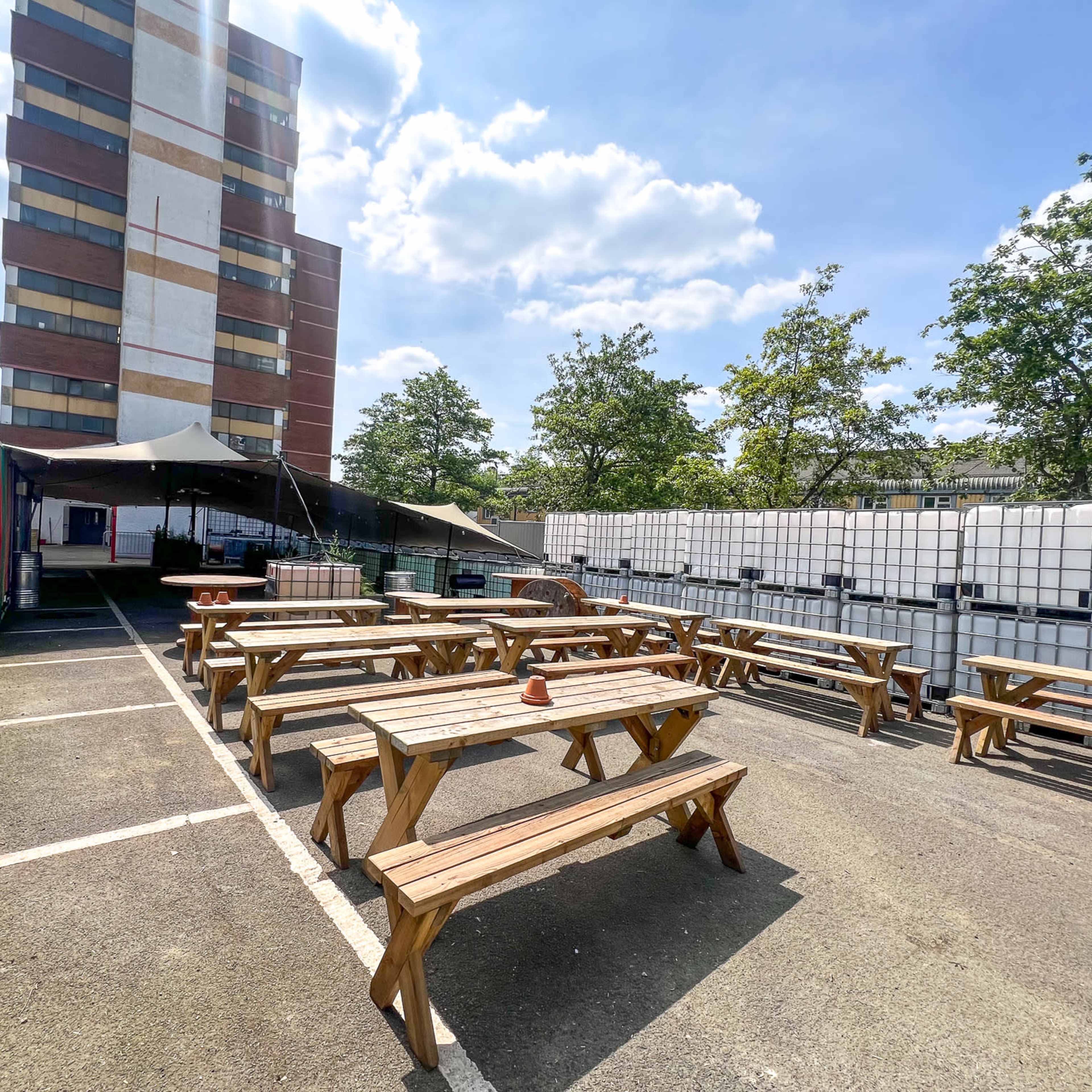 The scene features an outdoor seating area with wooden picnic tables surrounded by large water storage containers and a tall building in the background.
