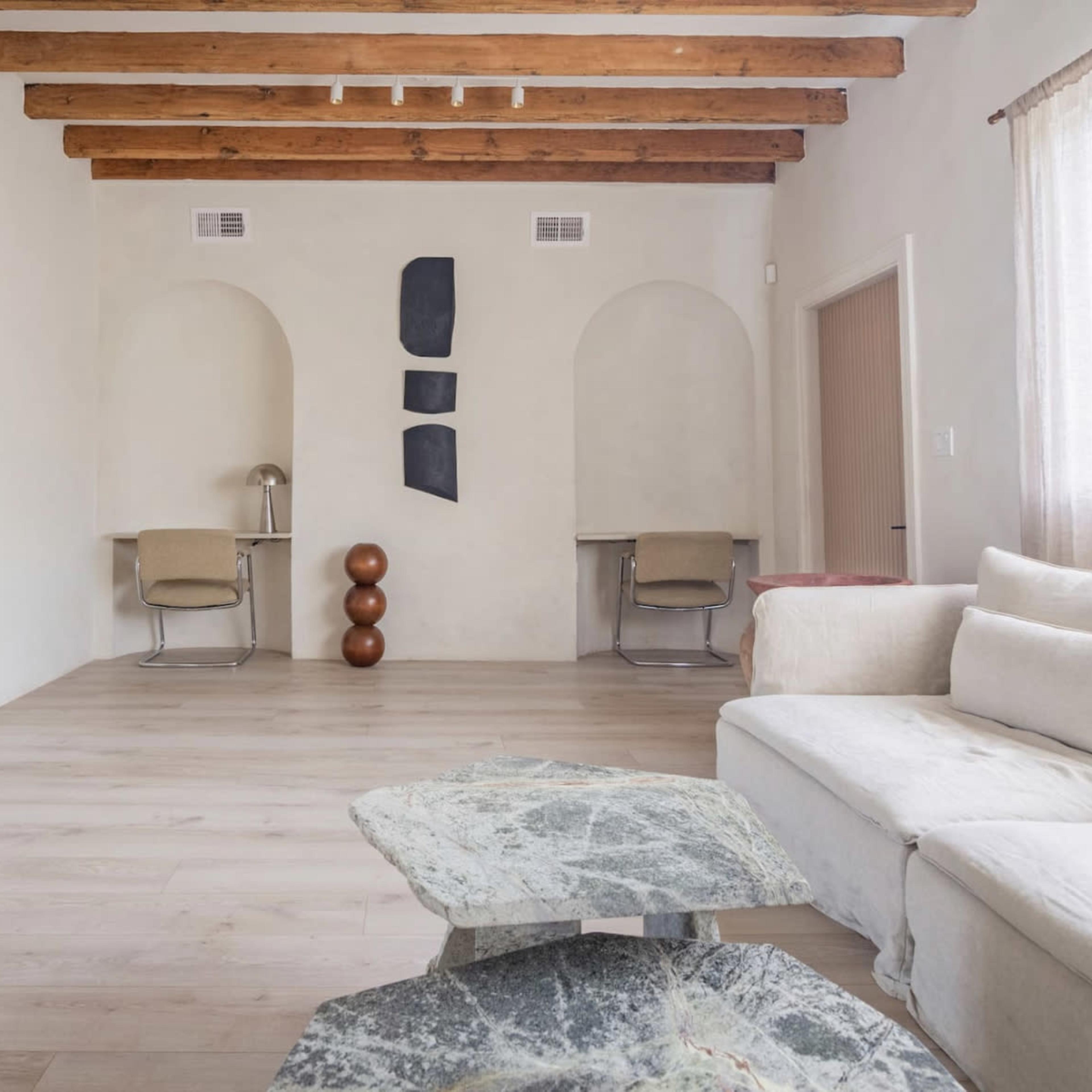 A spacious room with wooden beamed ceilings, featuring a modern sofa, two abstract wall decorations, and two work desks.