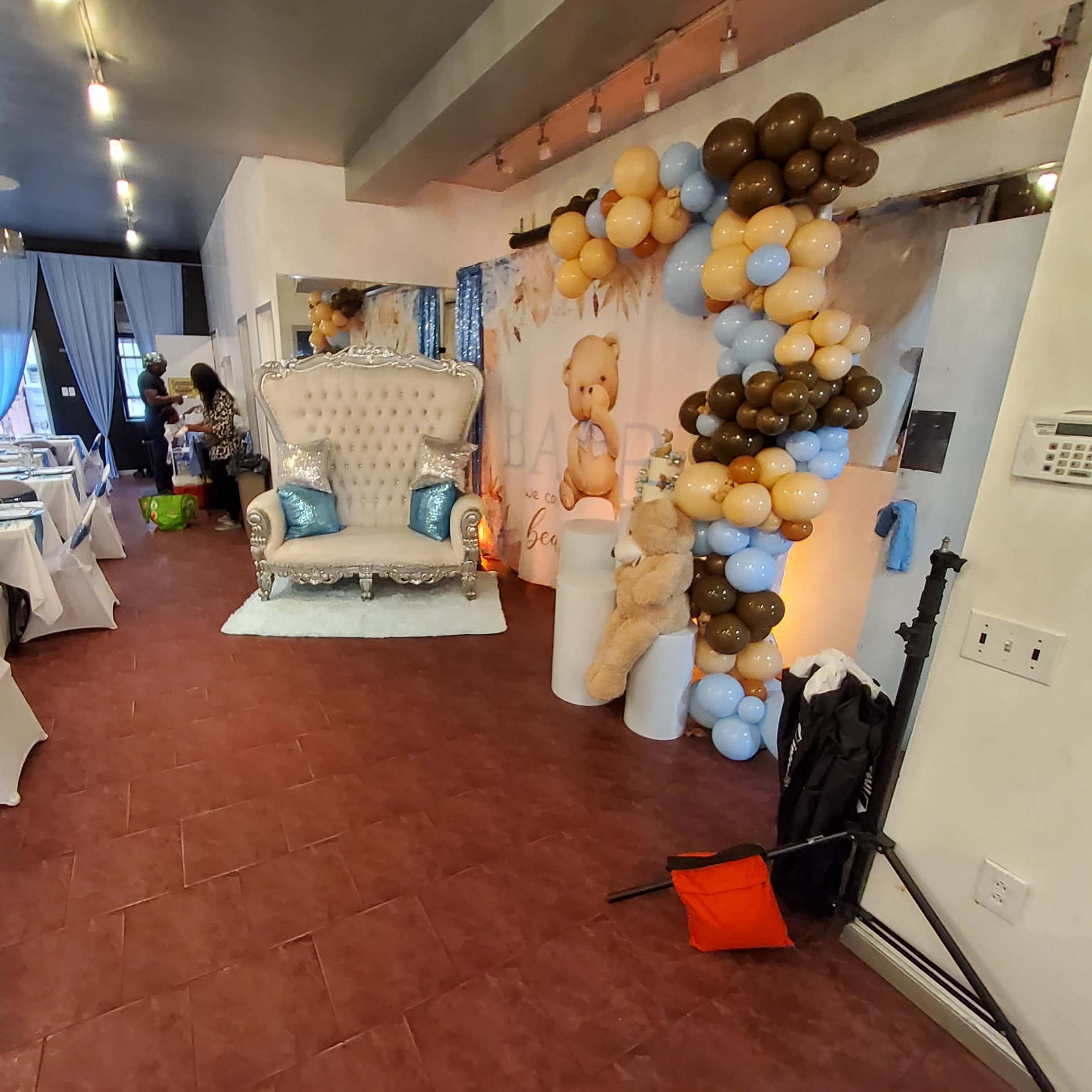 A decorated event space with tables set for a celebration, featuring a large white throne chair surrounded by colorful balloons and a backdrop with a teddy bear theme.