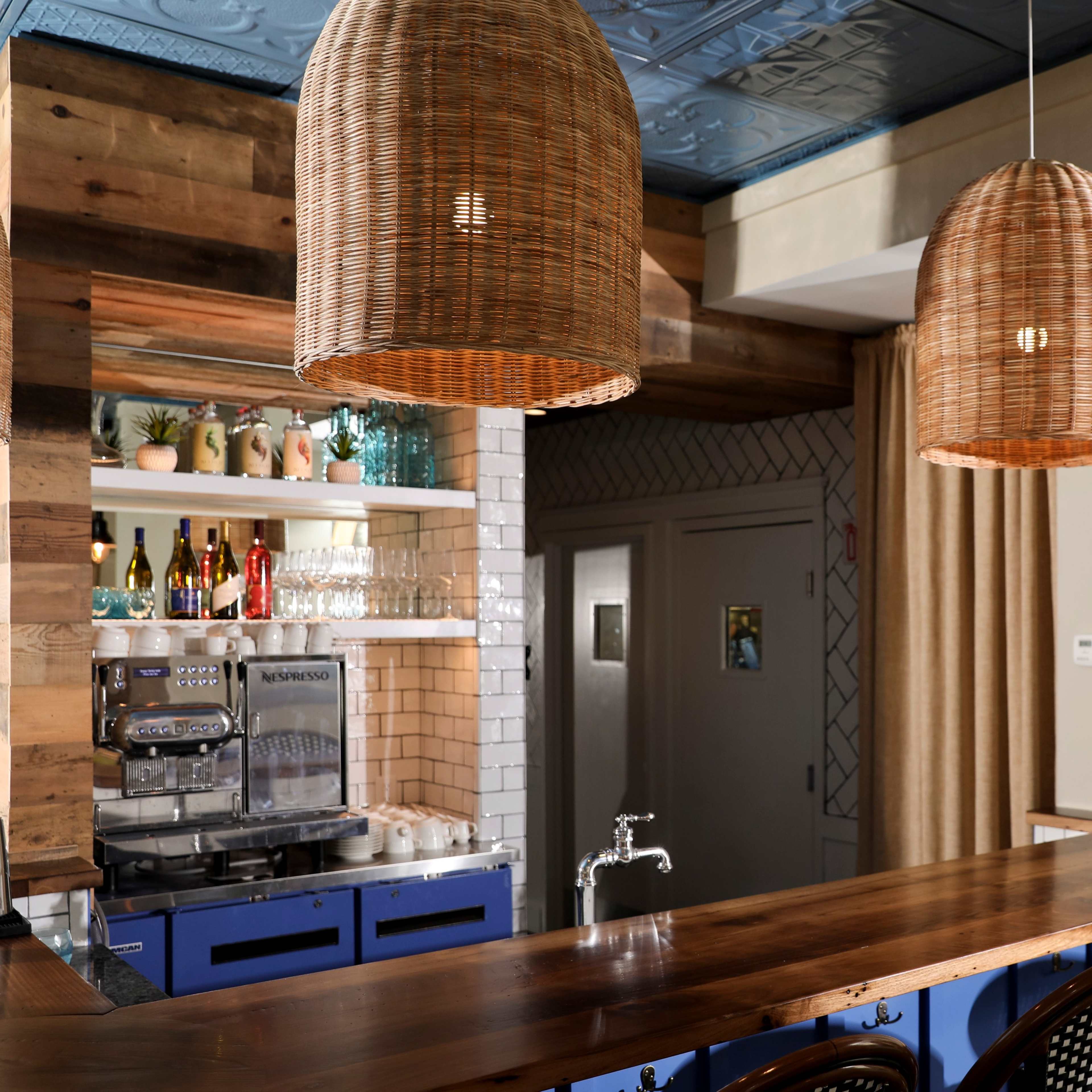 A bar area with a wooden countertop, woven pendant lights, and a shelf displaying various bottles and glassware.