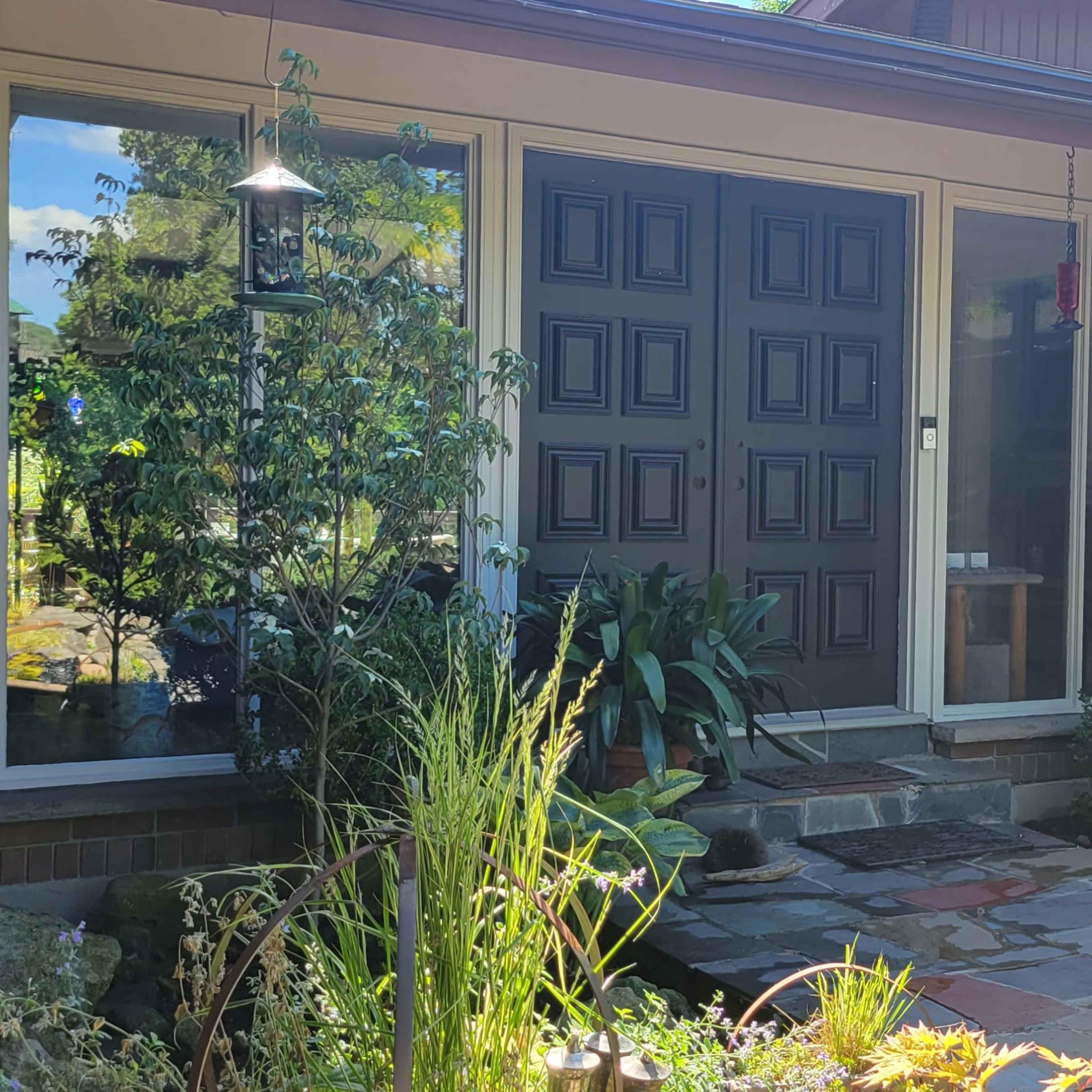 A front entrance of a house featuring double black doors, large windows, and a stone pathway surrounded by plants and landscaping.