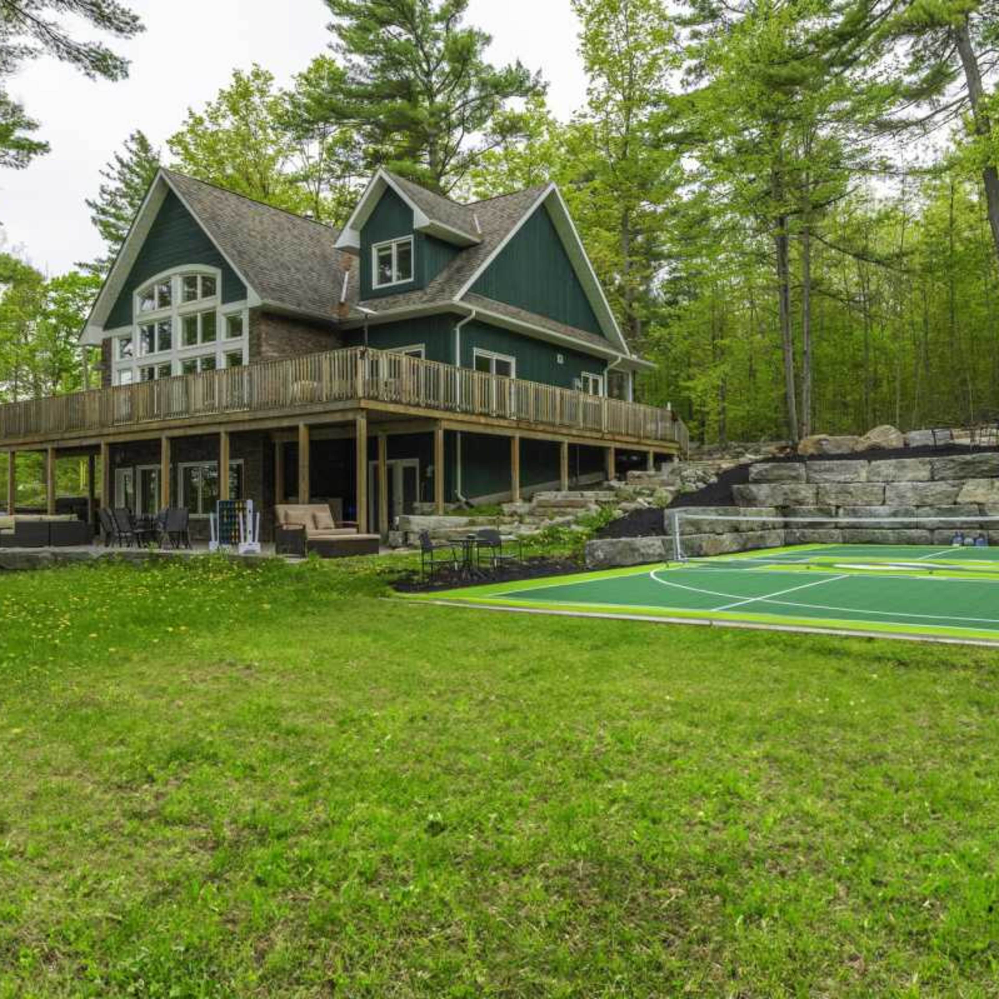 A large green and brown house with a deck overlooks a grass yard with a sports court and surrounding trees.