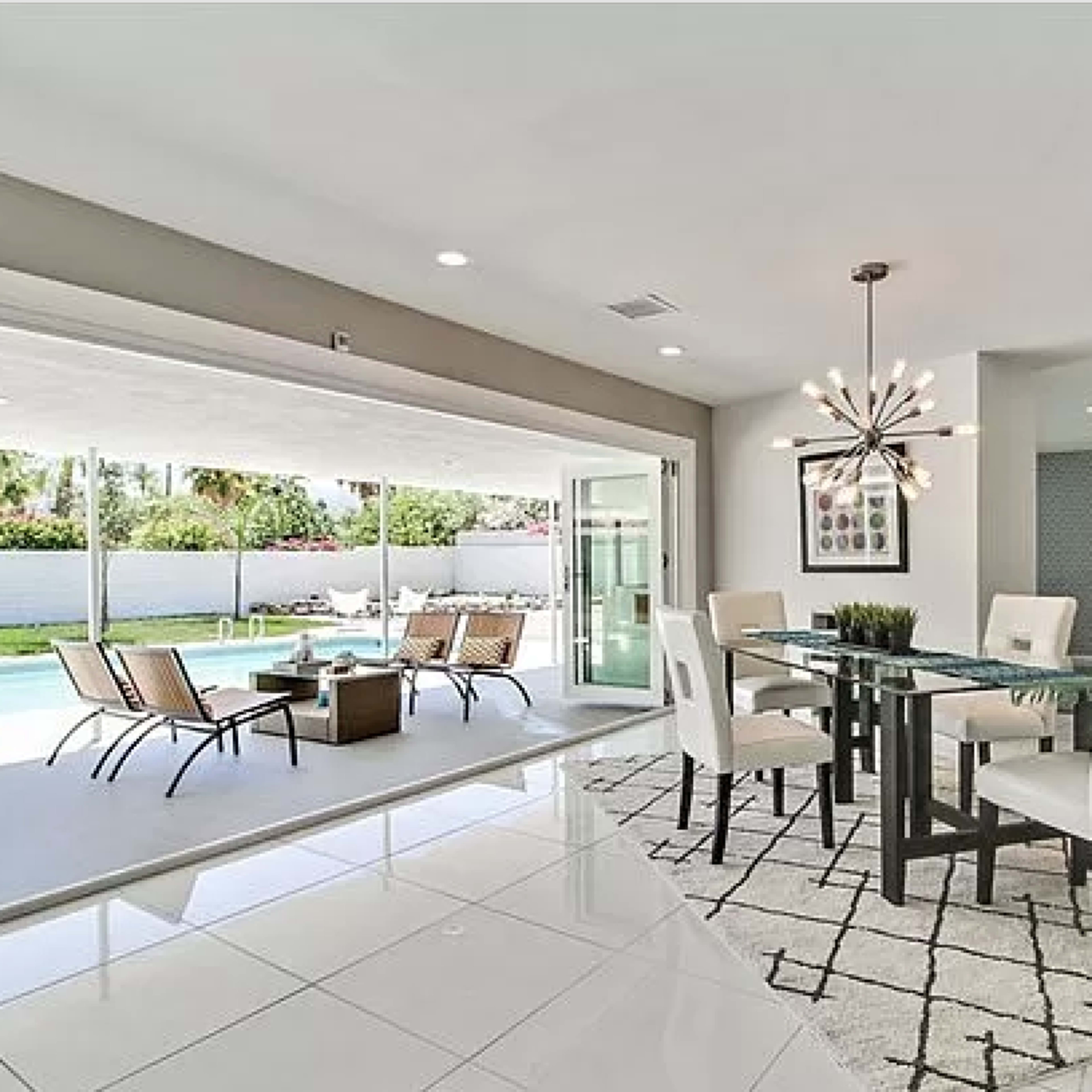 A modern dining area with a glass table and white chairs overlooks a pool and patio through large sliding doors.