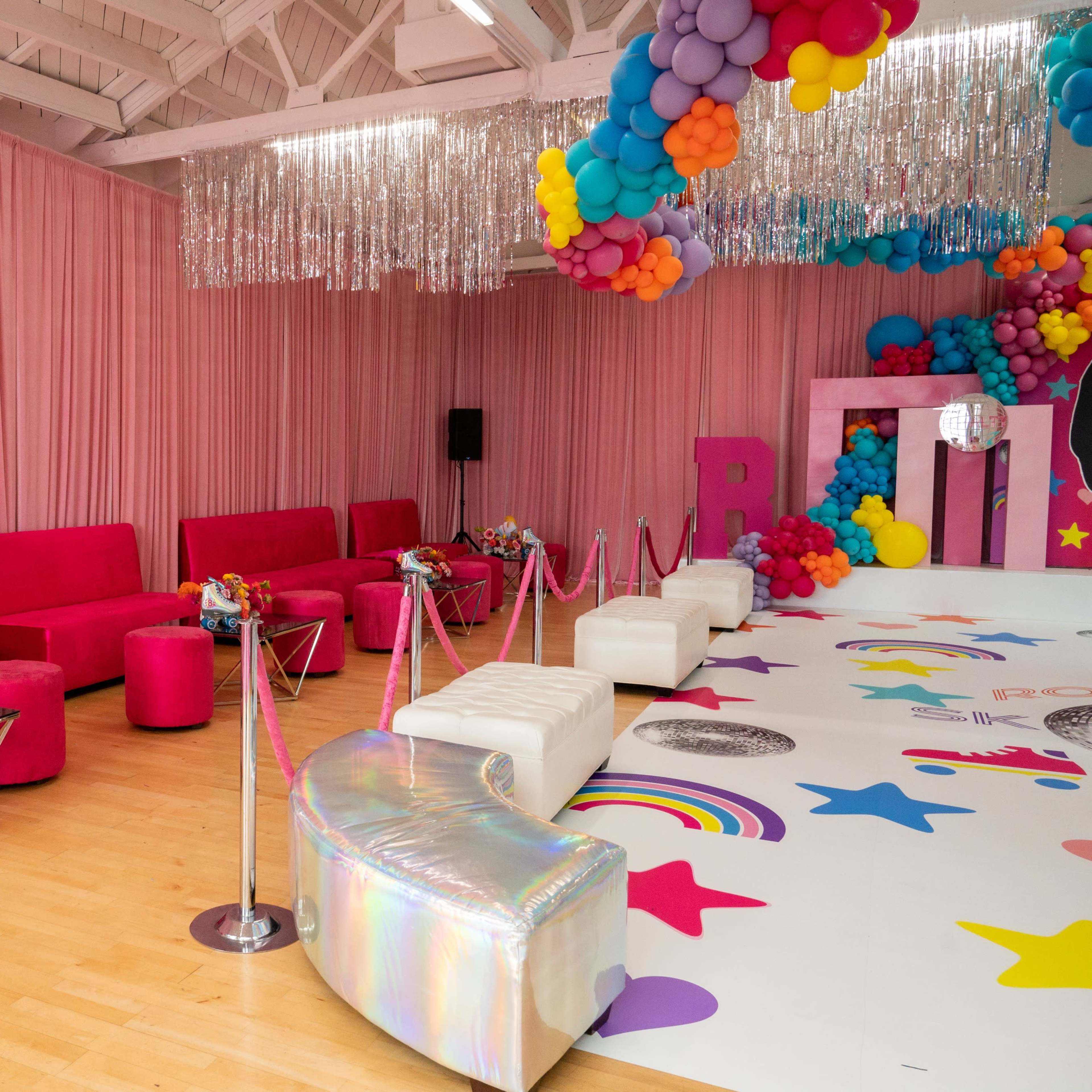 The scene features a brightly decorated event space with pink seating, colorful balloons, and a vibrant, star-patterned floor.