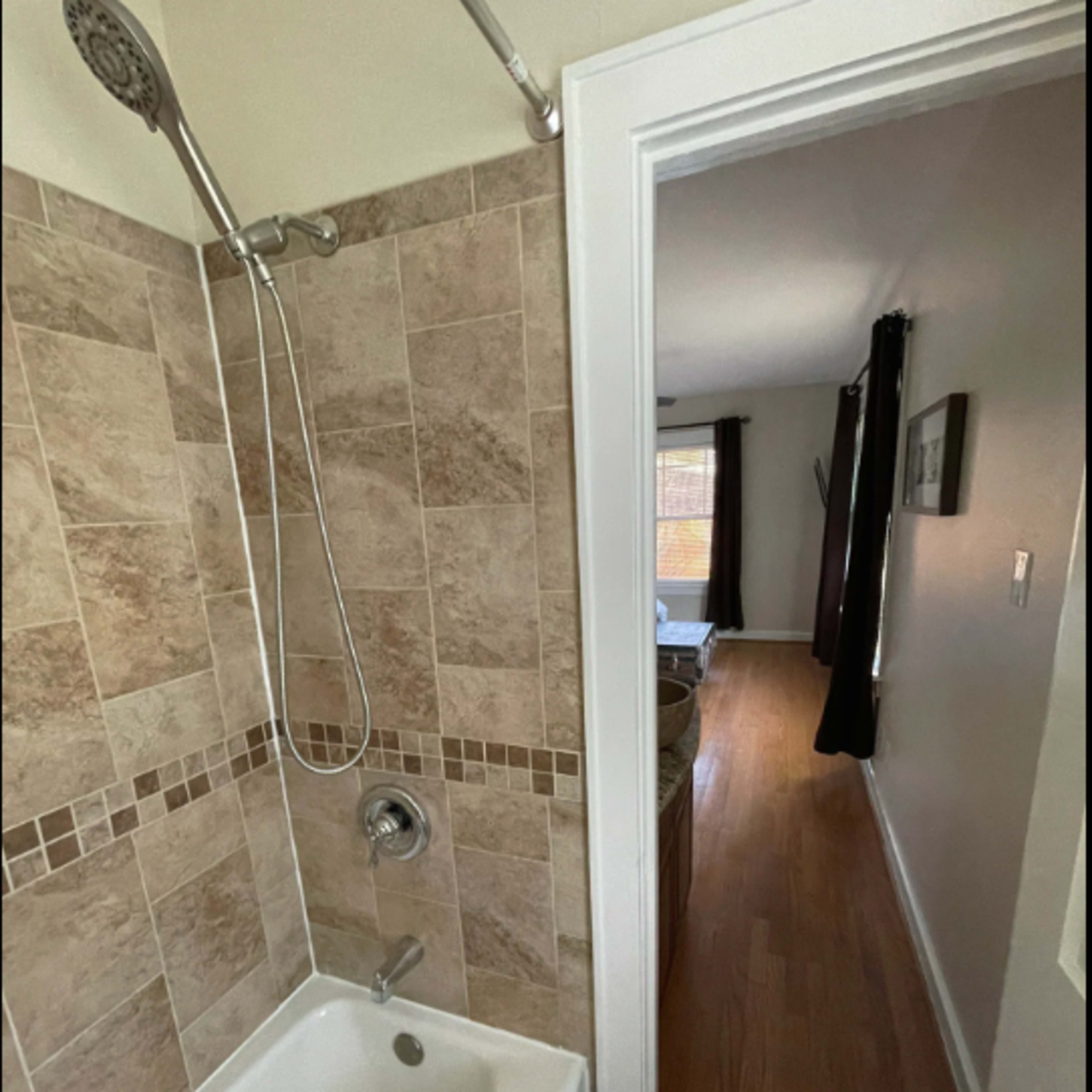 A bathroom with a shower and bathtub combination, featuring tiled walls and a doorway leading to a hallway with wooden floors.