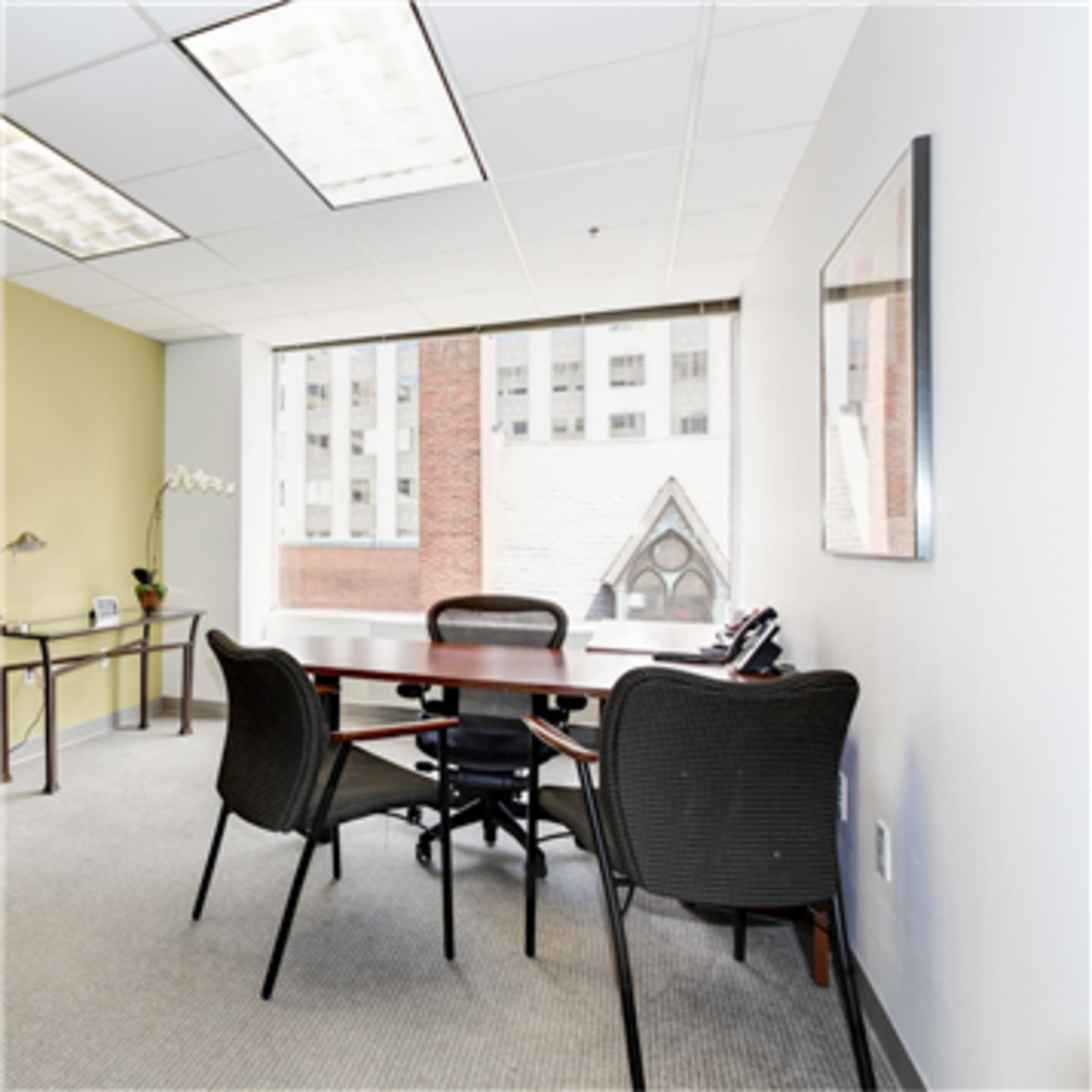 An office space with a round table and four chairs, a phone on the table, and a large window overlooking a brick building.
