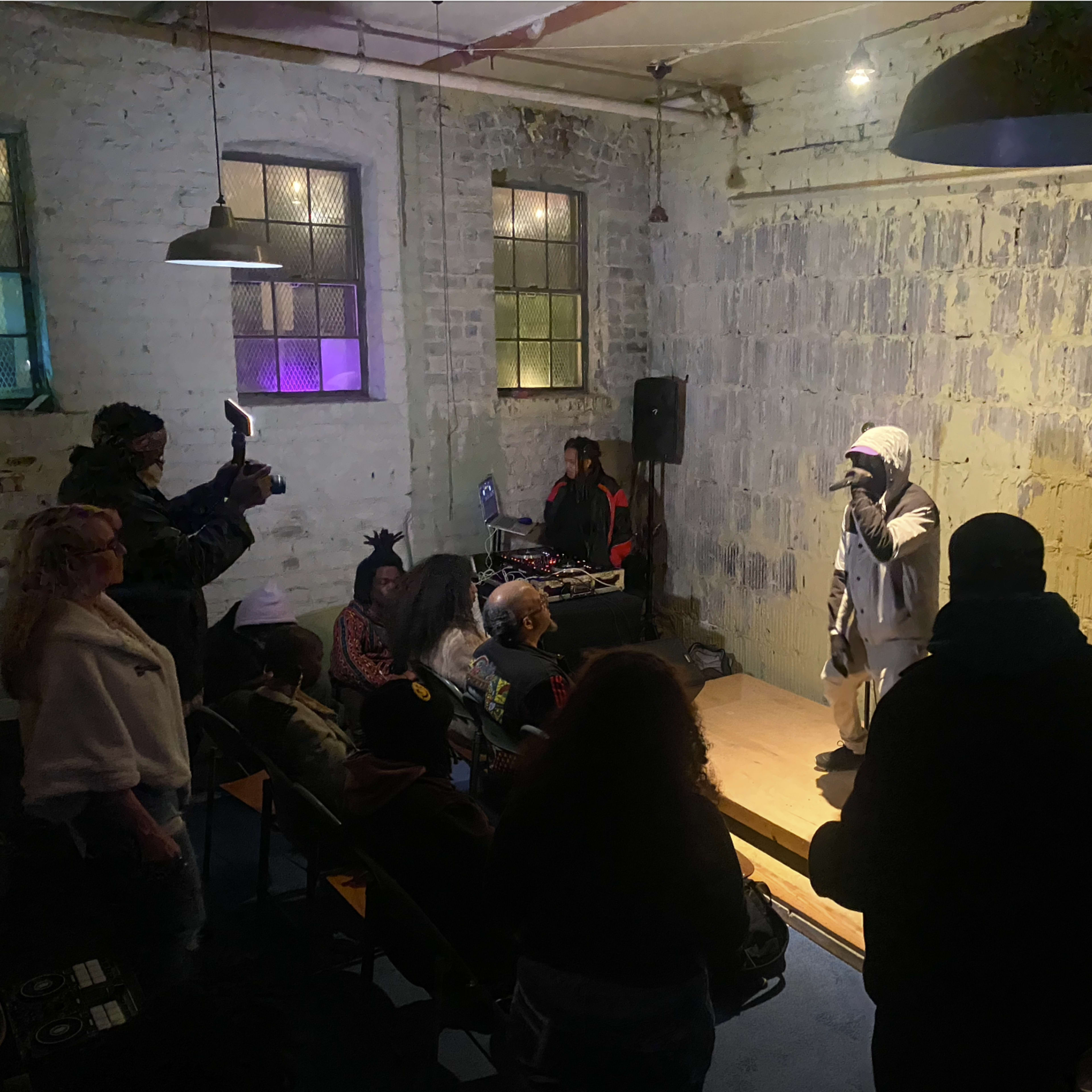 A performer is on stage in a small, dimly lit venue while an audience watches and a cameraman records the scene.