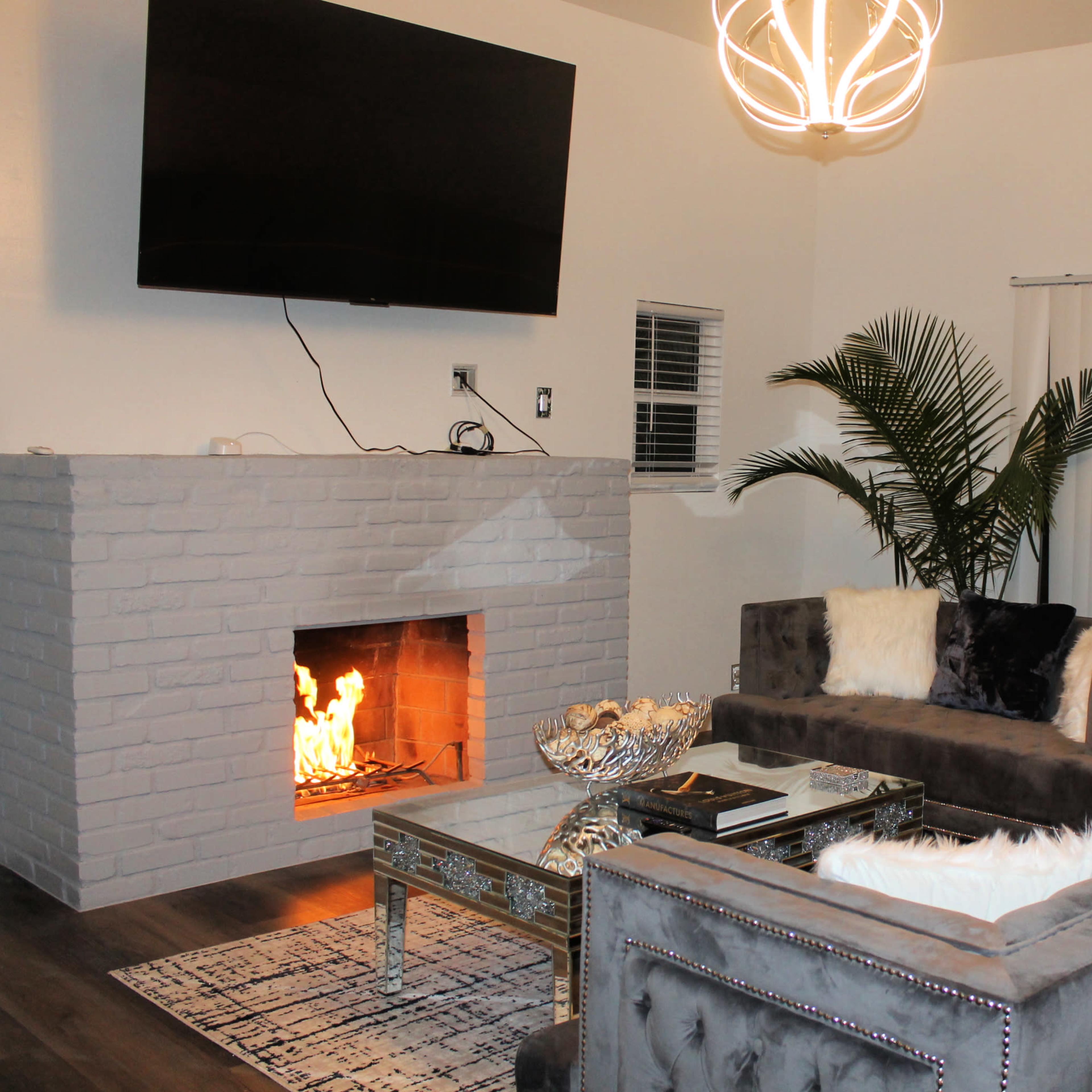 A modern living room features a gray sectional sofa, a glass coffee table, a brick fireplace with a flame, and a wall-mounted television, complemented by a tall indoor plant and vertical blinds.